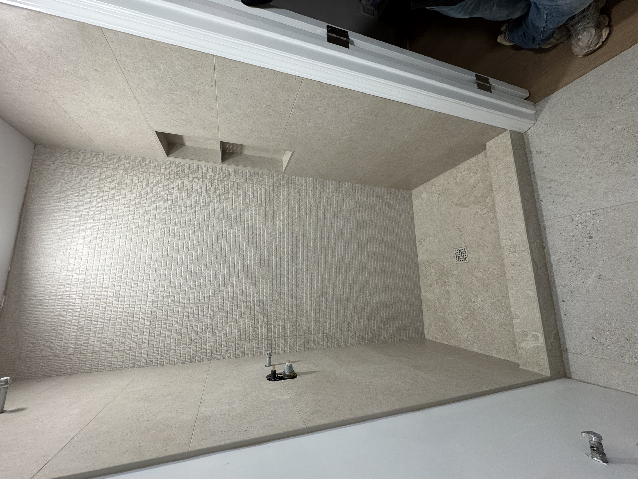 Empty shower area with beige tiles on the wall and floor, featuring a drain and water supply fixtures, and an alcove for toiletries, under construction or renovation.