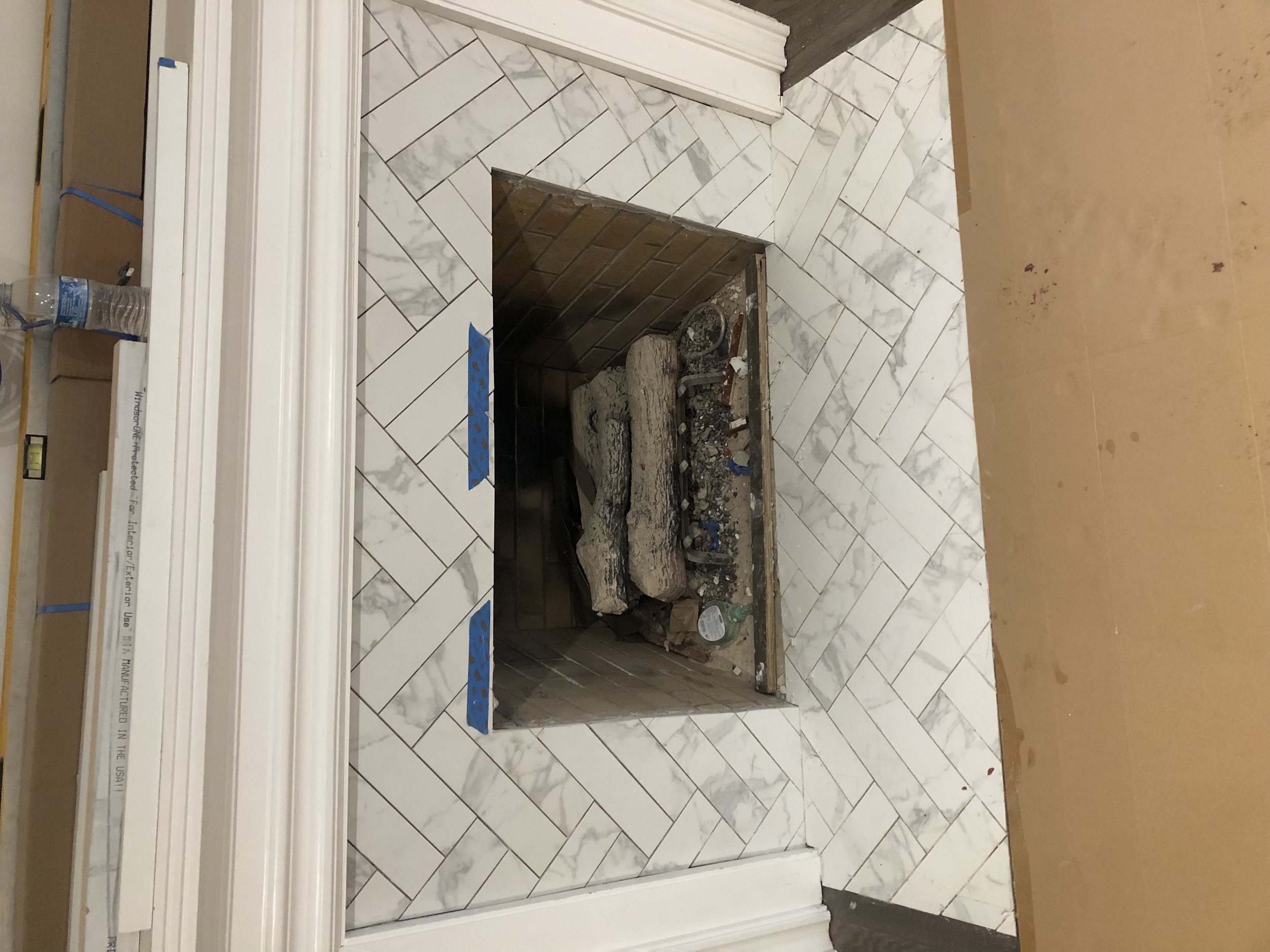 A fireplace with a white marble tile surround in a room under construction. The interior of the fireplace is unfinished with visible bricks and construction debris.