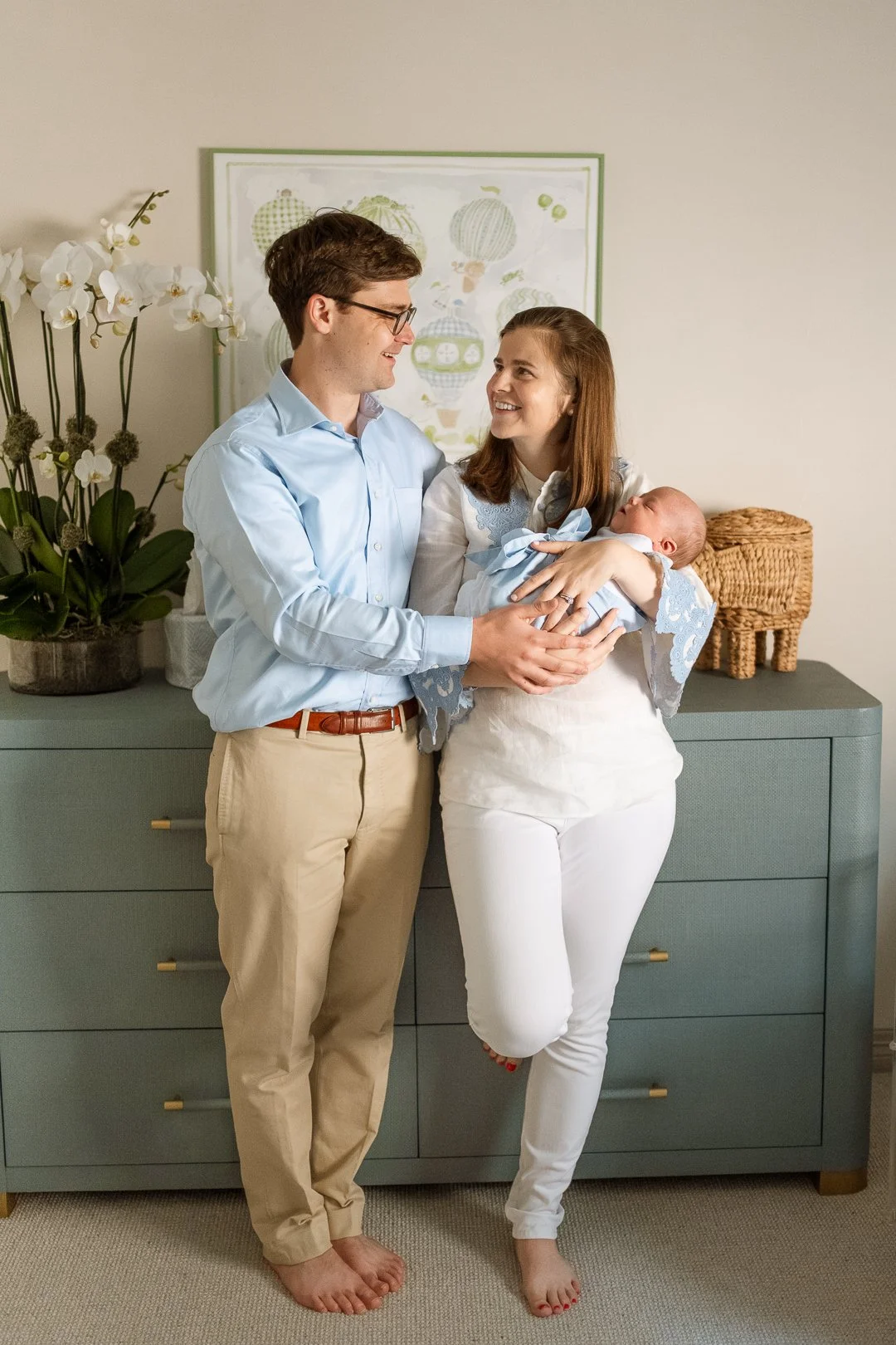 A happy couple holding a newborn baby in a cozy room with a dresser, orchid plant, and framed artwork.