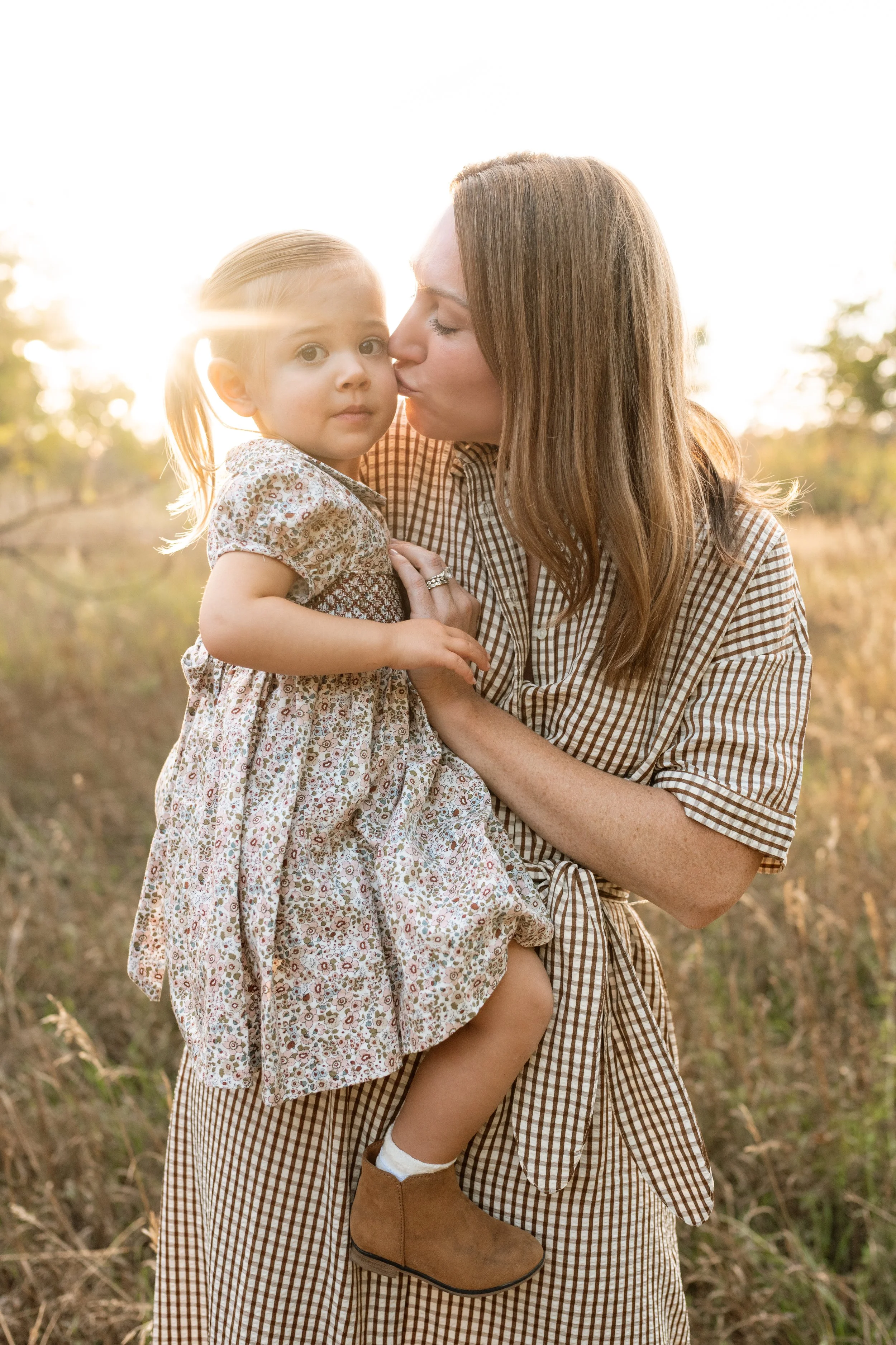 A woman holding a young girl outdoors at sunset, with the woman kissing the girl on the cheek.