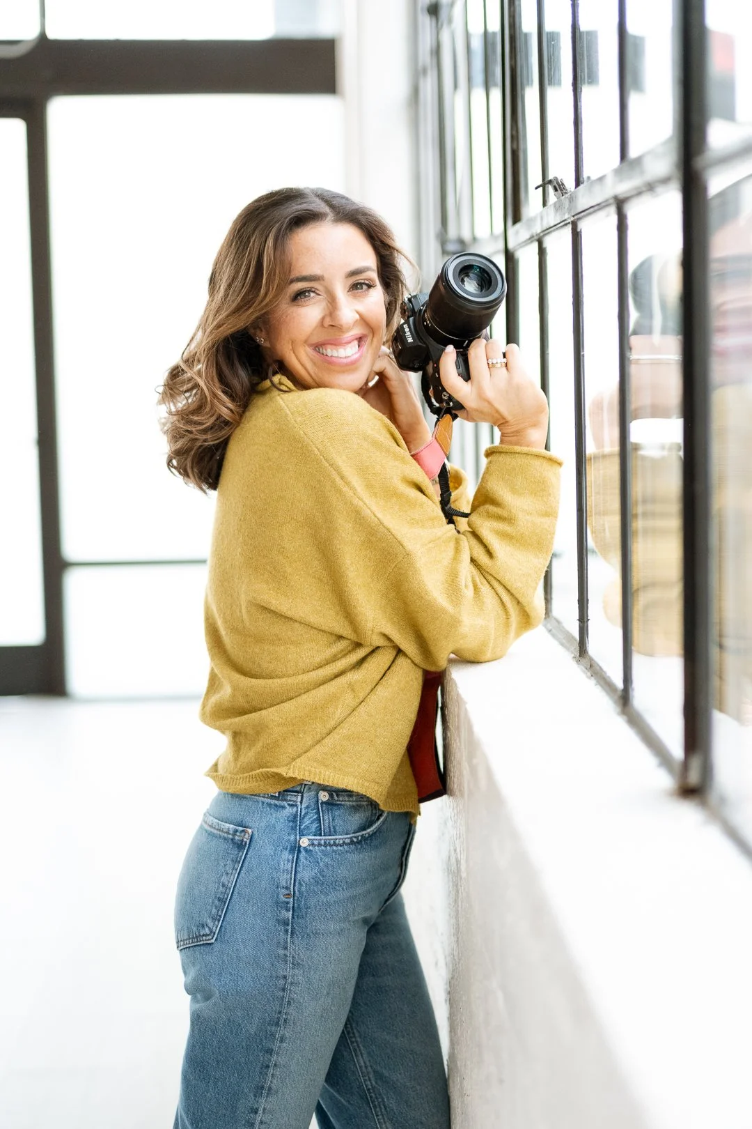A young woman with wavy brown hair, wearing a yellow sweater and blue jeans, holding a camera while standing by a large window and smiling.