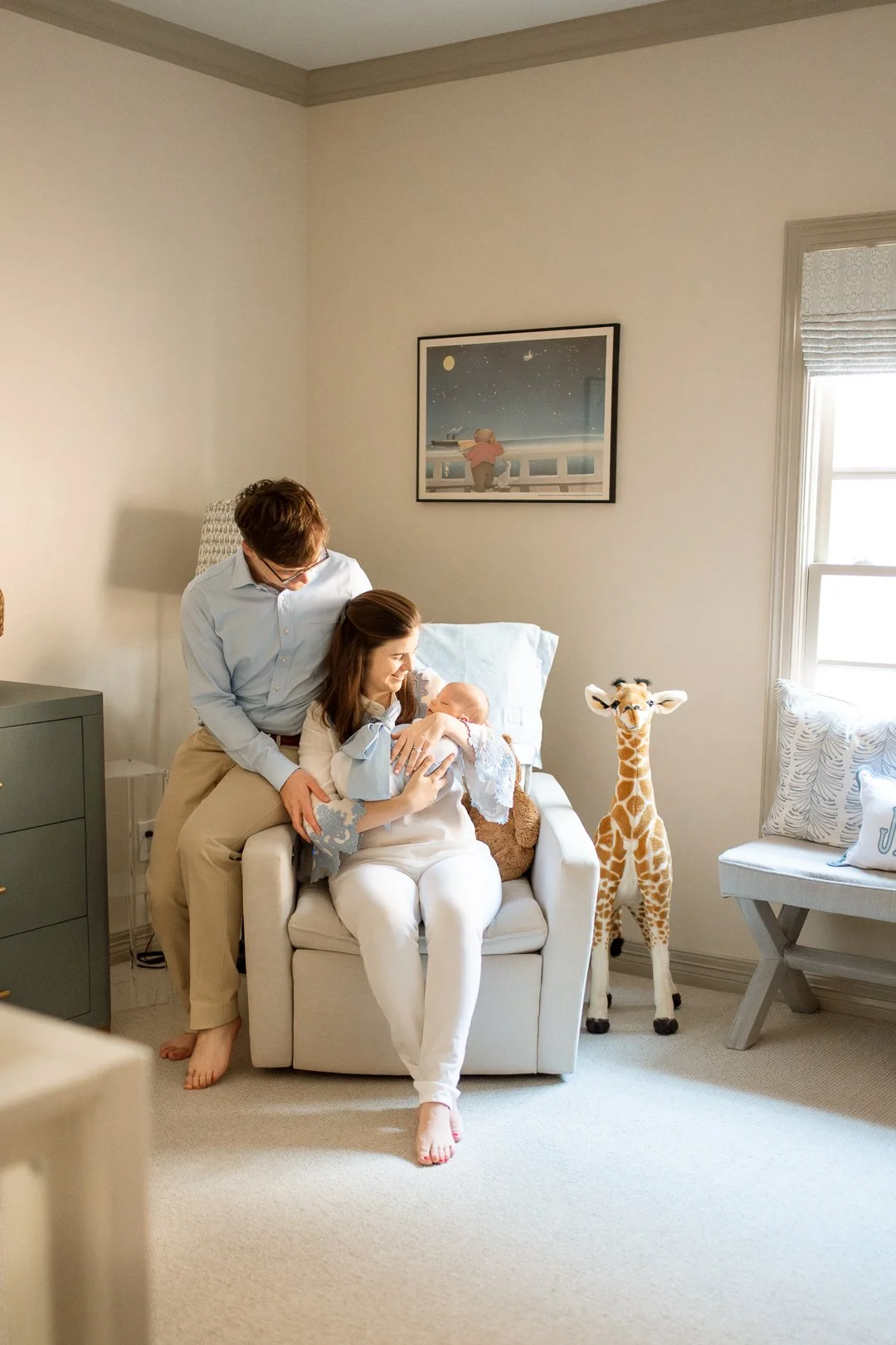 A woman holding a newborn in a nursery, with a man standing beside her, both smiling. There is a stuffed giraffe and a pillow on a bench by the window, and a framed picture on the wall.
