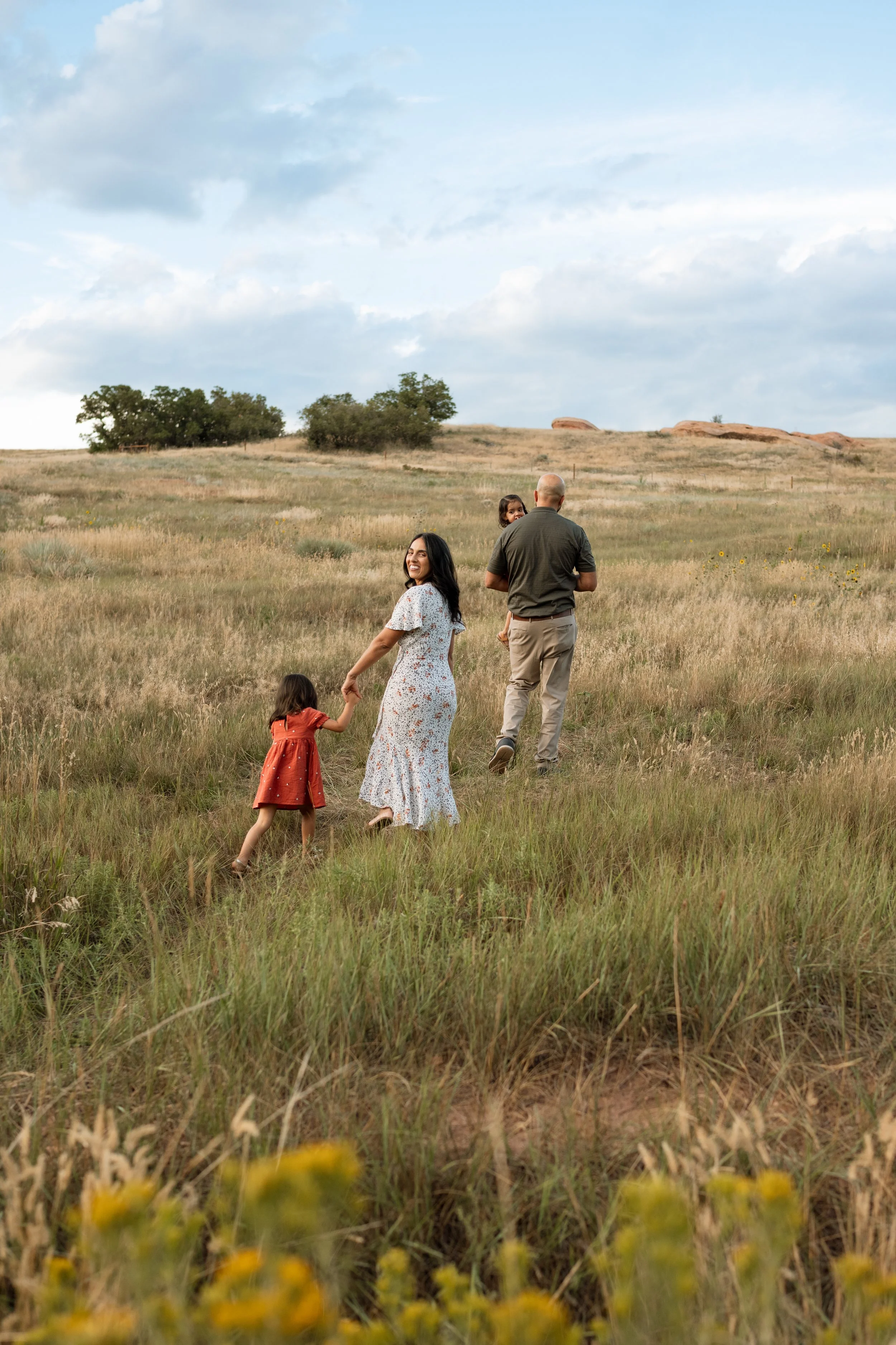 A family walking through a grassy field, with two adults and two young girls, one of whom is carried by the man.