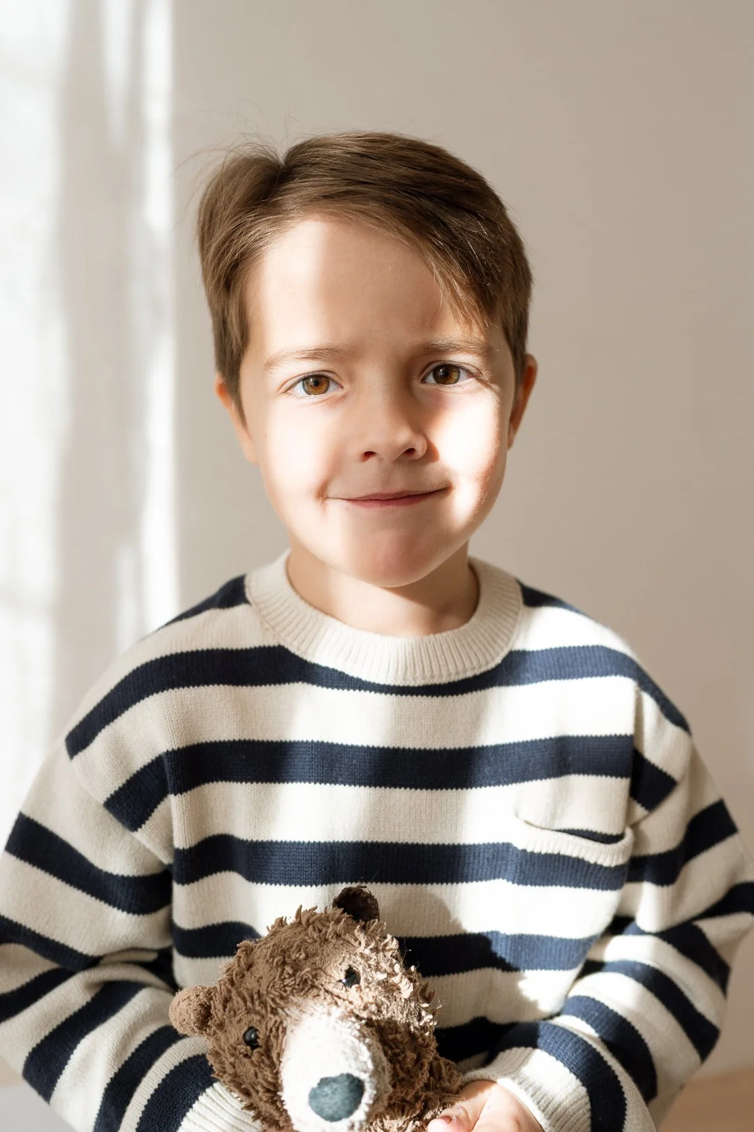 A young boy holding a plush brown teddy bear, wearing a white sweater with navy blue stripes, standing near a xwhite curtain with sunlight.