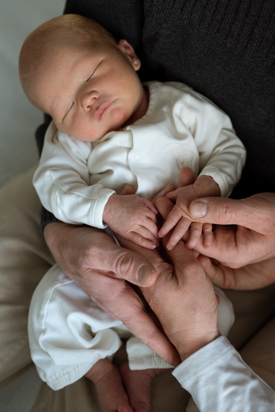 A newborn baby sleeping, being held gently by two adult hands, with the baby's tiny hand grasping an adult finger.