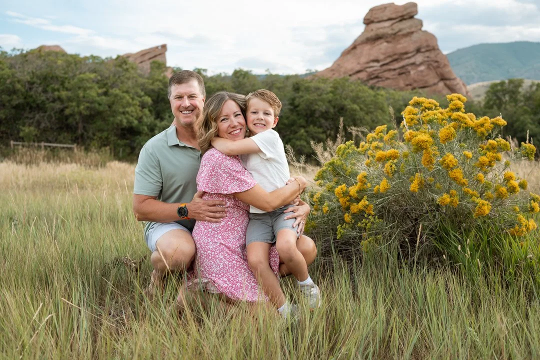 A happy family of three, including a man, woman, and boy, posing outdoors in a grassy field with rock formations and trees in the background.