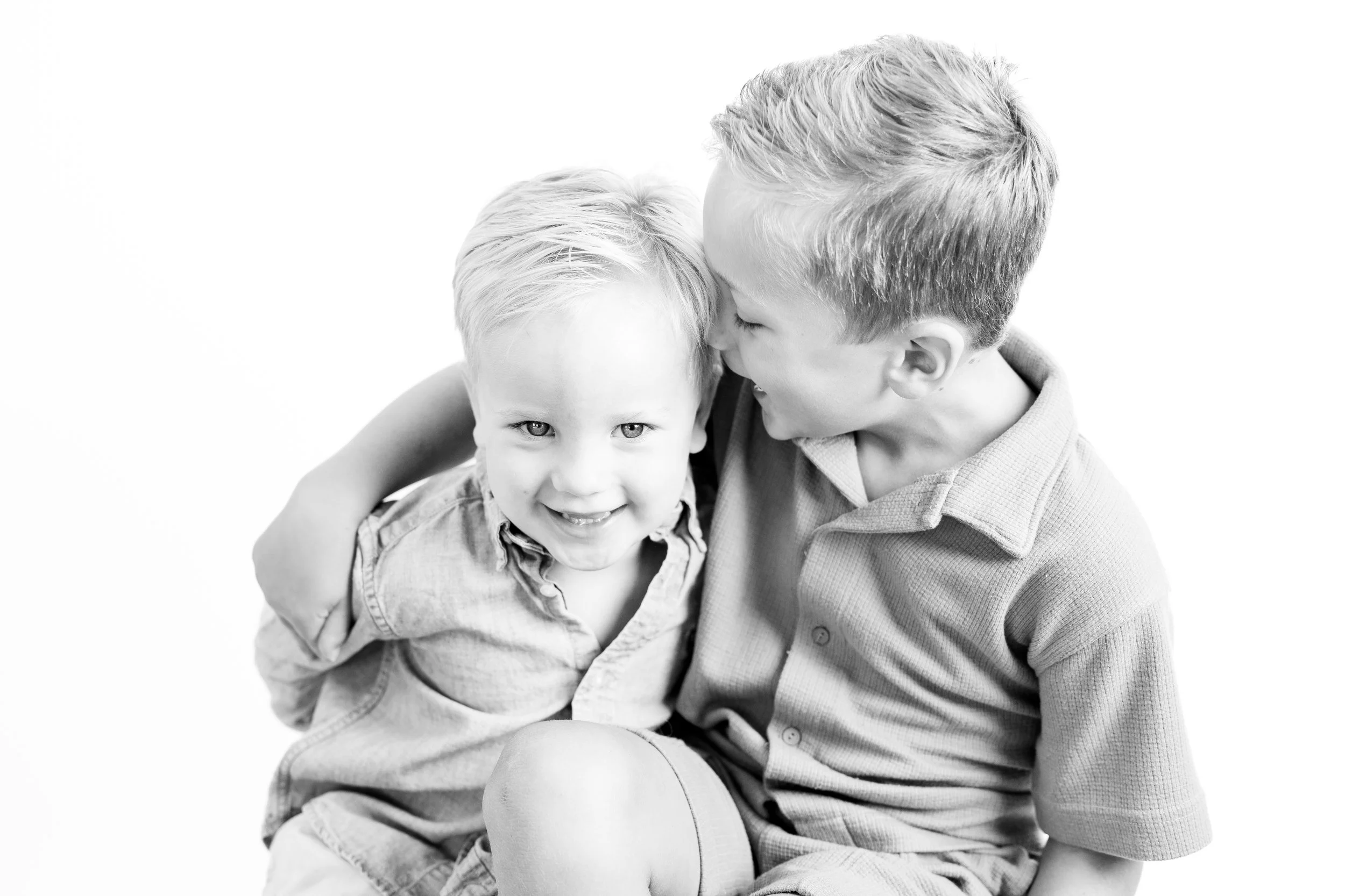 Two young boys sitting closely, one laughing and the other whispering or speaking into his ear, in a black and white photo.