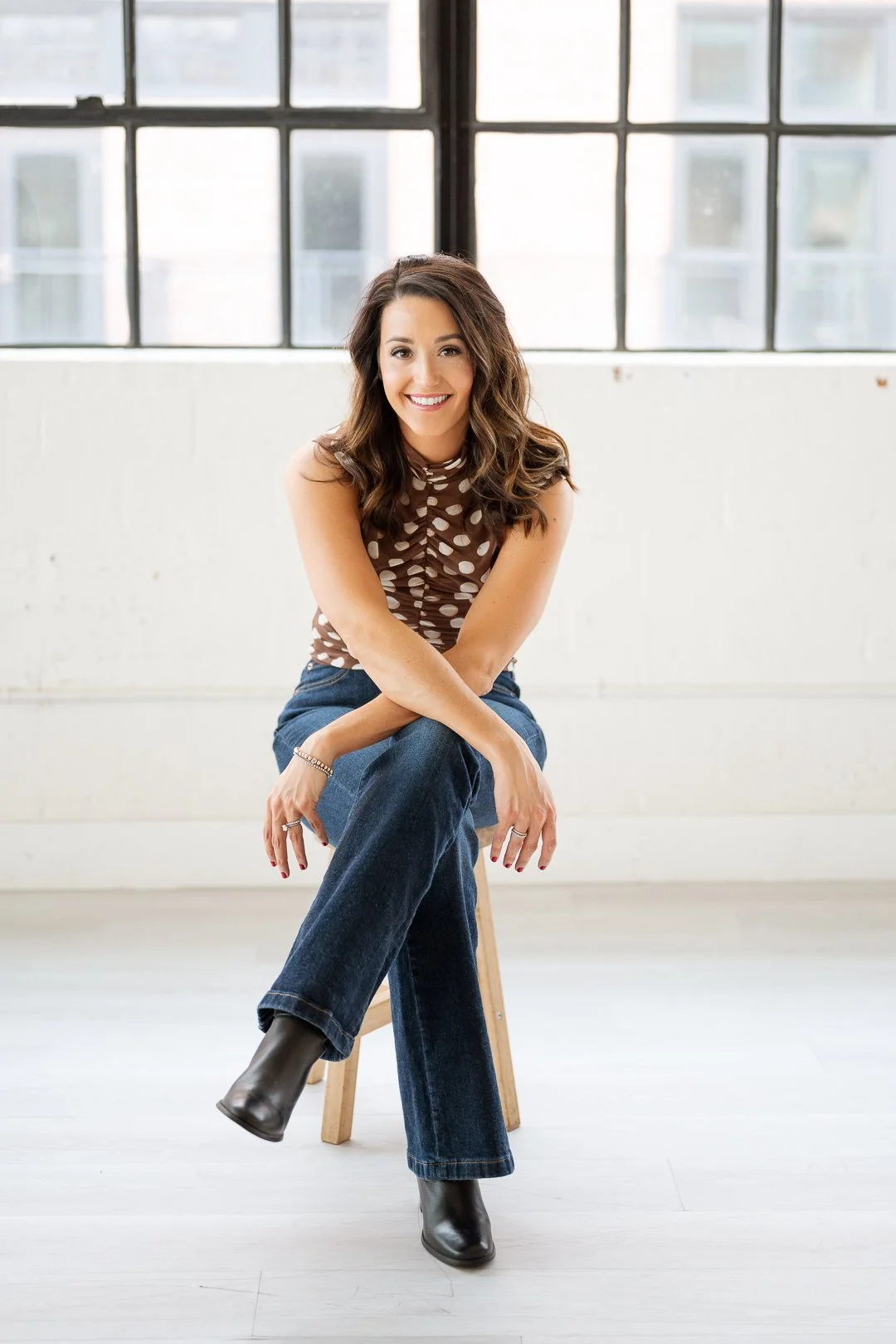 A woman with wavy brown hair smiling, sitting on a stool in front of large windows in a bright, minimalist room, wearing a brown polka-dot sleeveless top, blue jeans, and black boots.