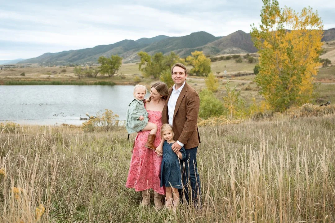 A family of four standing in a grassy field near a lake with mountains and trees in the background during fall.