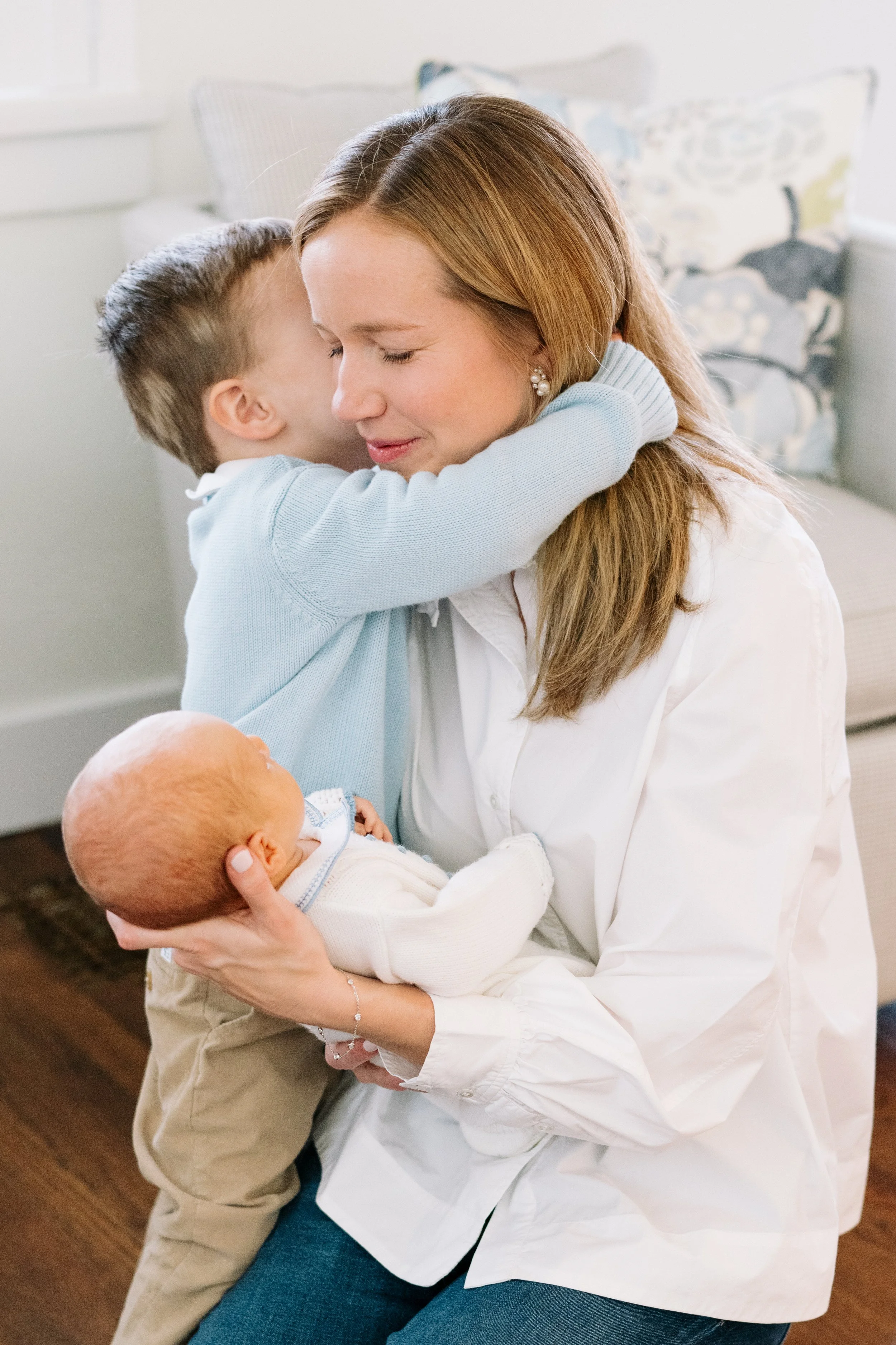 A woman holding a newborn and a young boy hugging her tightly, with a neutral-colored couch in the background.