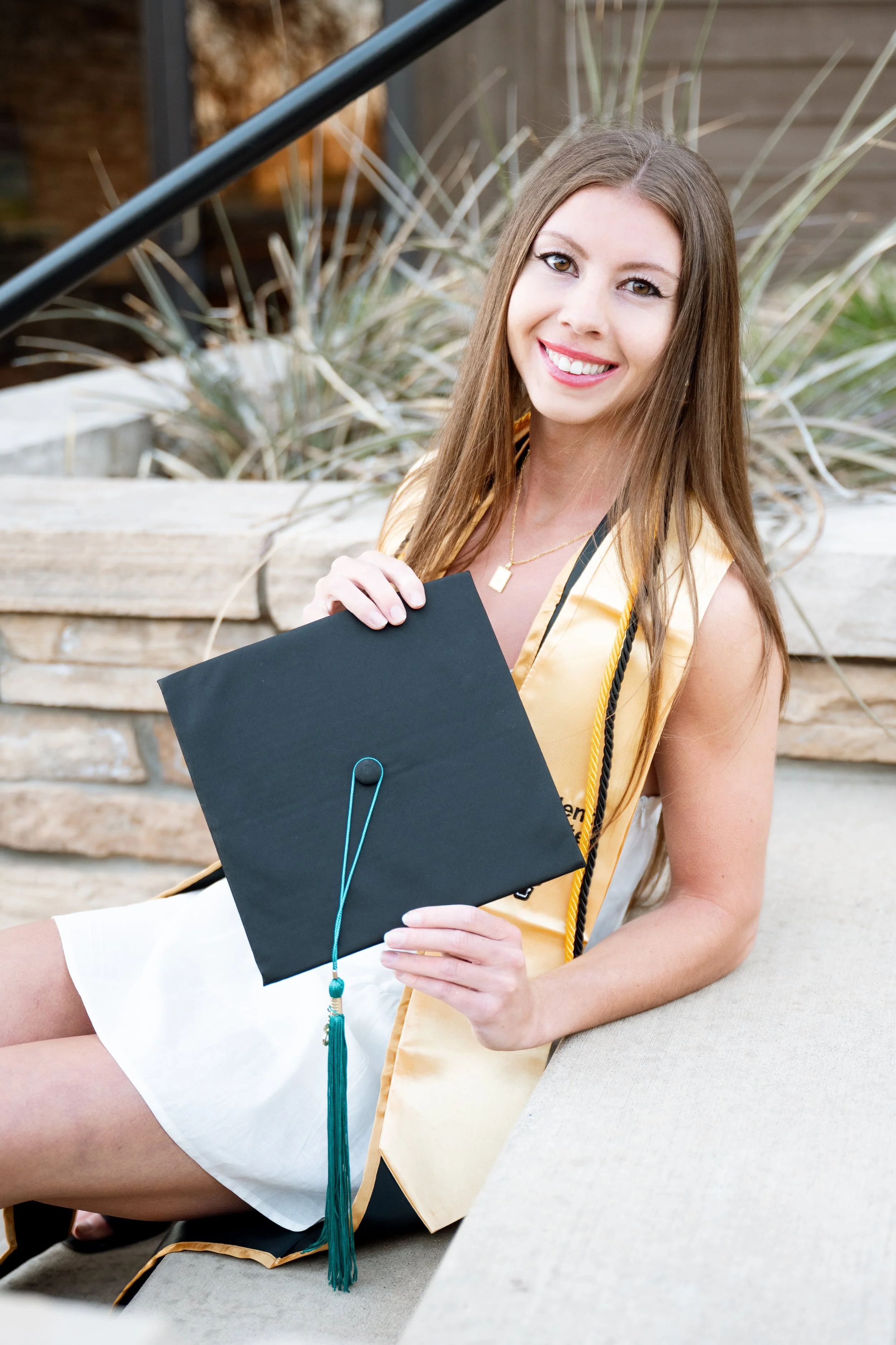 A young woman in a yellow graduation gown sitting outdoors, holding a black graduation cap with a blue tassel, smiling at the camera.