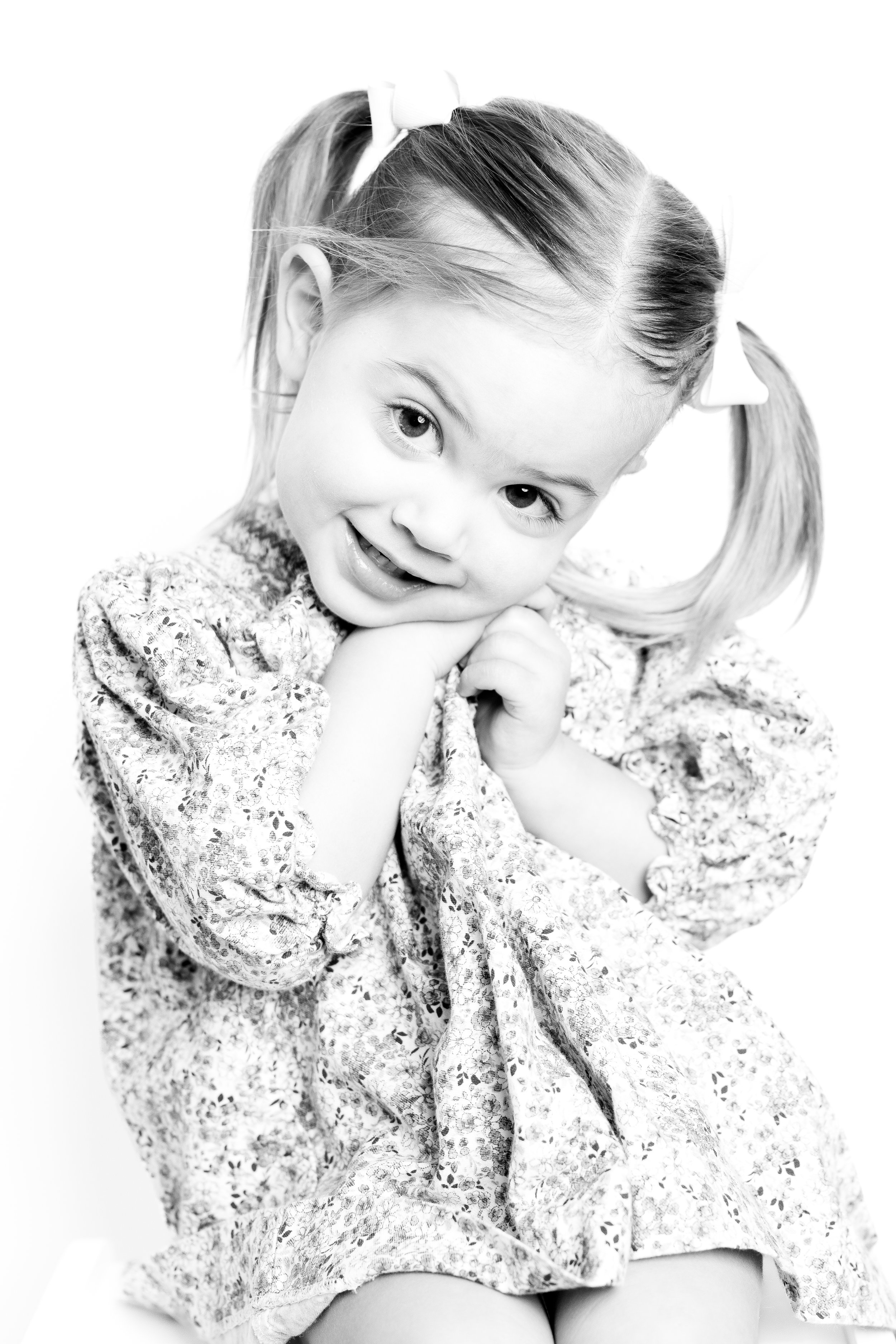 A young girl with pigtails, wearing a floral dress, smiling and resting her chin on her hands in a black-and-white photograph.