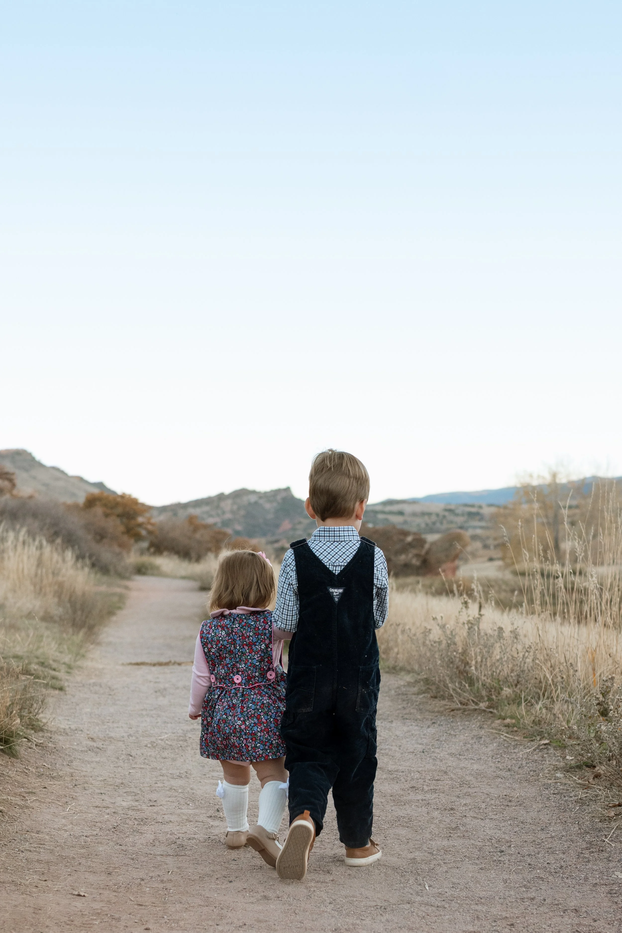 Two children, a boy and a girl, walk hand-in-hand along a dirt path through a dry, grassy landscape with hills in the distance.
