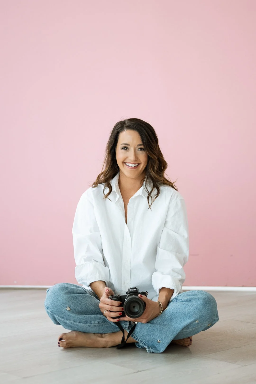 A woman with brown hair, wearing a white shirt and blue jeans, sitting on the floor with a camera, smiling at the camera against a pink wall.