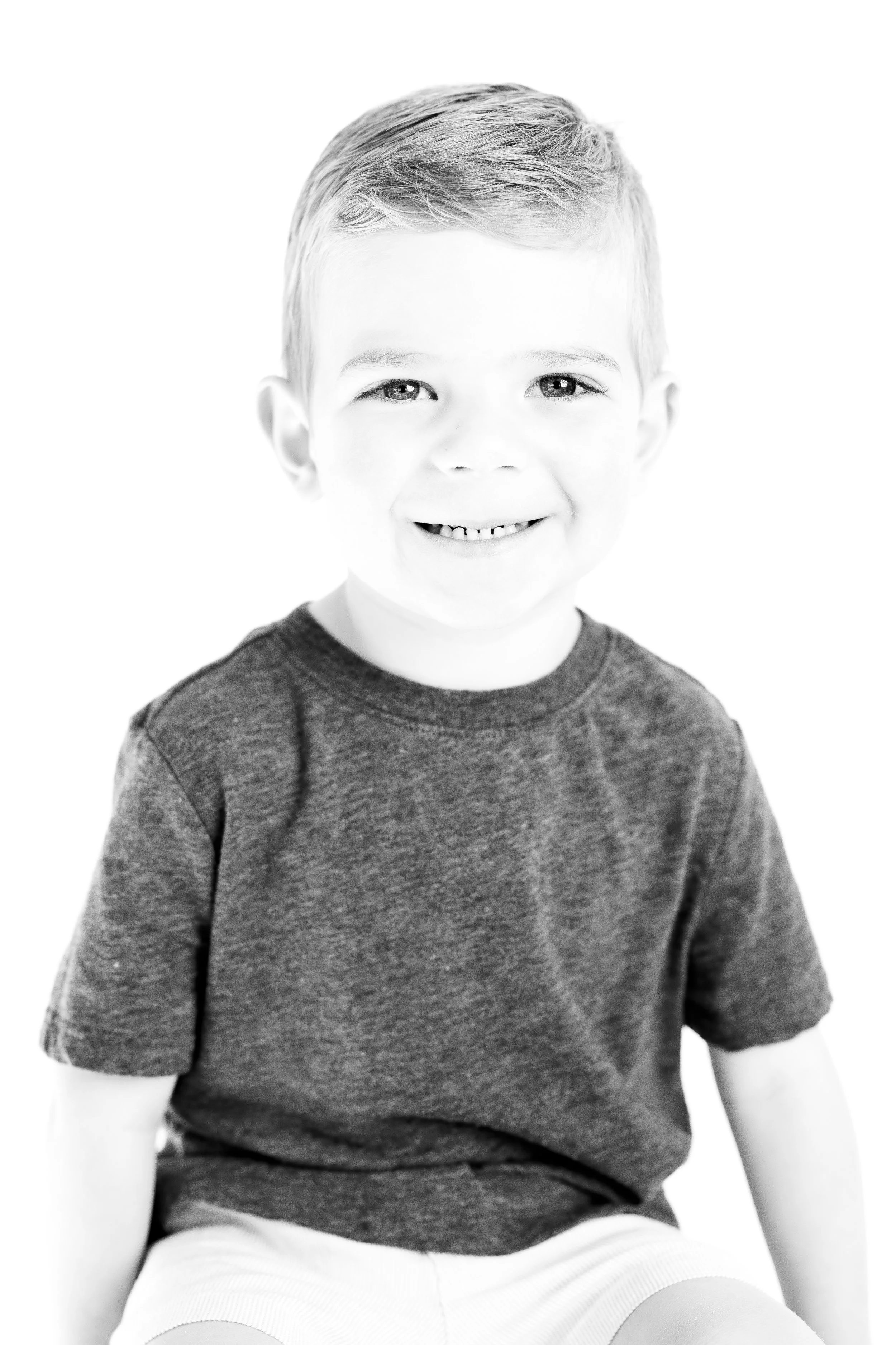 A young boy with short hair smiling, wearing a dark t-shirt and light-colored shorts, in black and white photography.