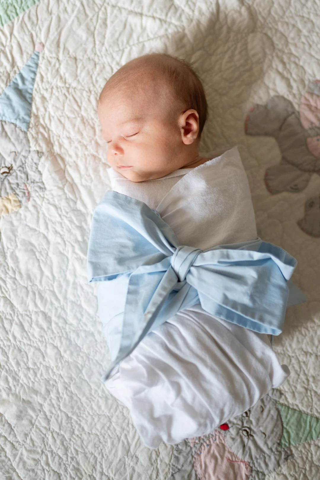 Sleeping baby wrapped in a white cloth with a large light blue bow, lying on a quilted blanket.