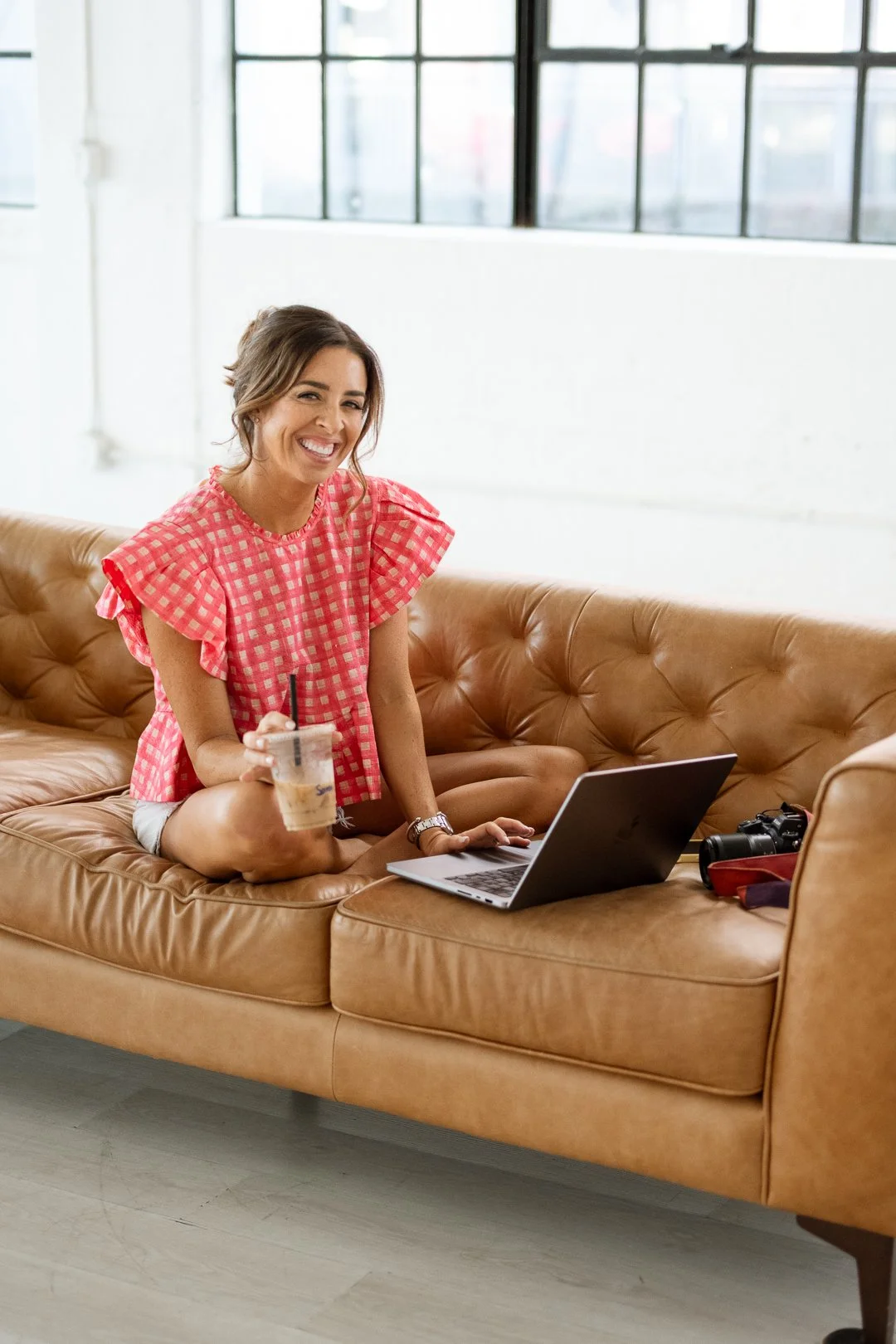 A woman sitting on a tan leather couch in a bright, industrial-style room with large windows. She is smiling, holding a drink, with a laptop, camera, and bag beside her.