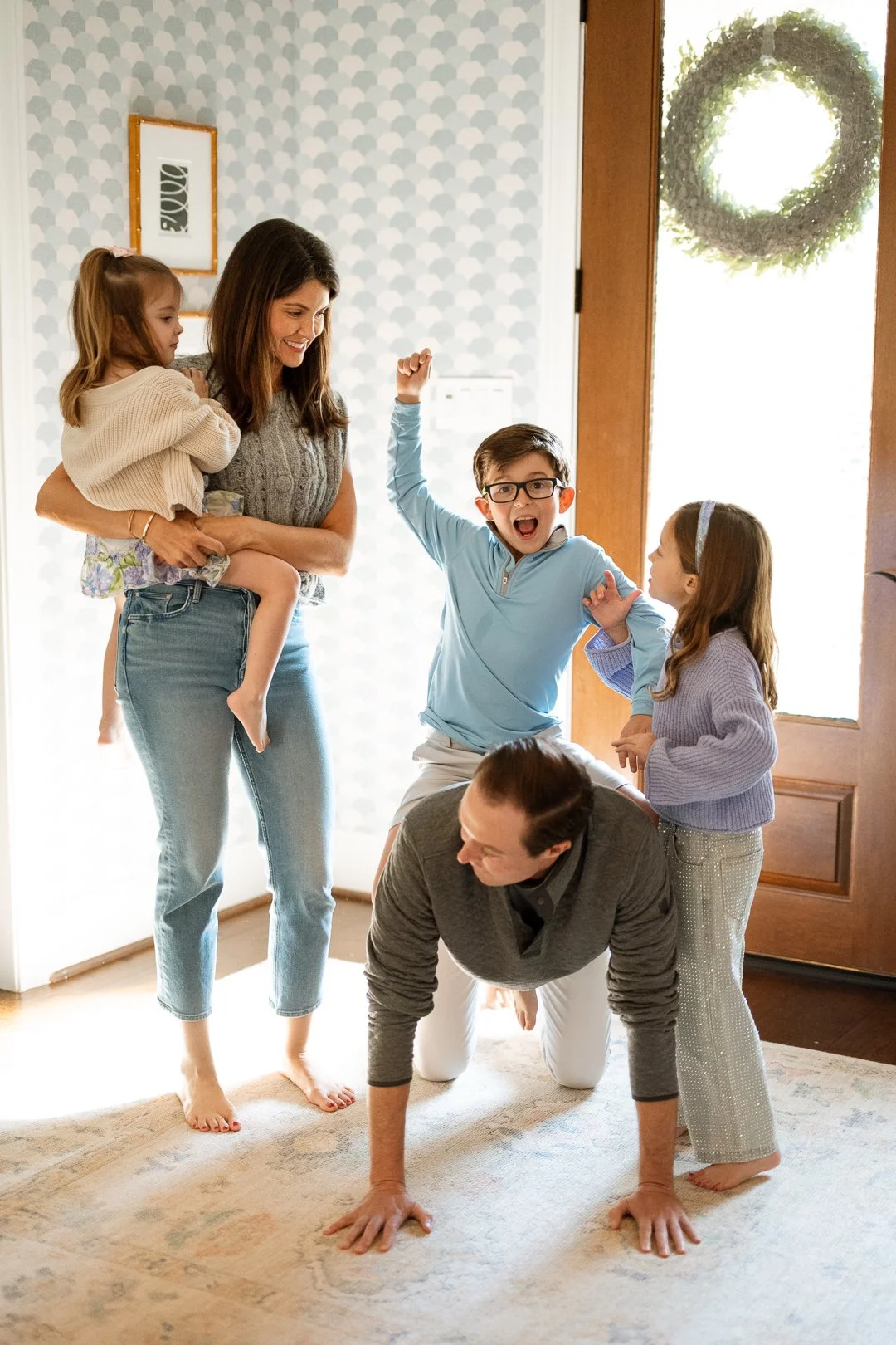 A family playing a game indoors in front of a door with a wreath. The woman is carrying a young girl, while two children, a boy with glasses and a girl, are engaged in a game with a man on all fours. The children look excited and happy.