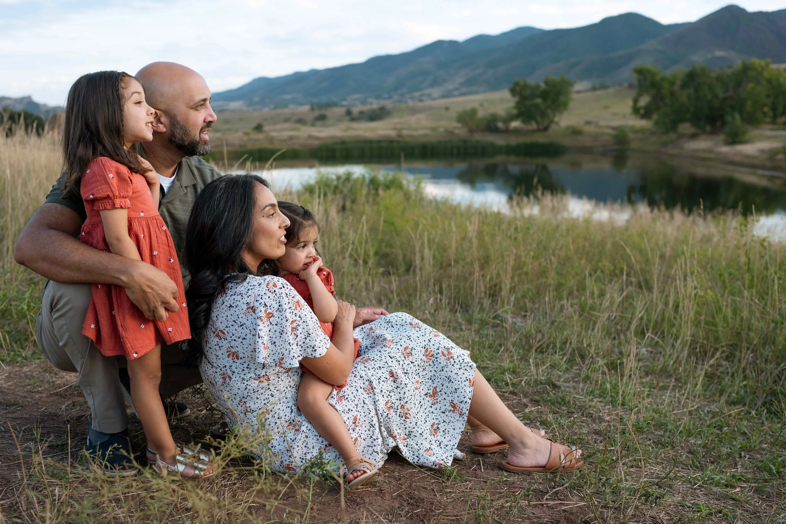 A family of five outdoors near a lake and mountains, sitting and standing on grass, enjoying a scenic view.