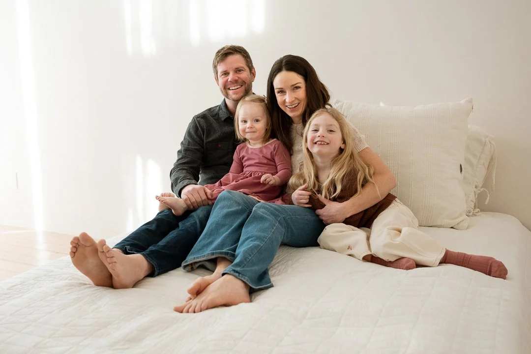 A happy family of four, including a father, mother, and two young daughters, sitting together on a bed in a bright room, smiling at the camera.
