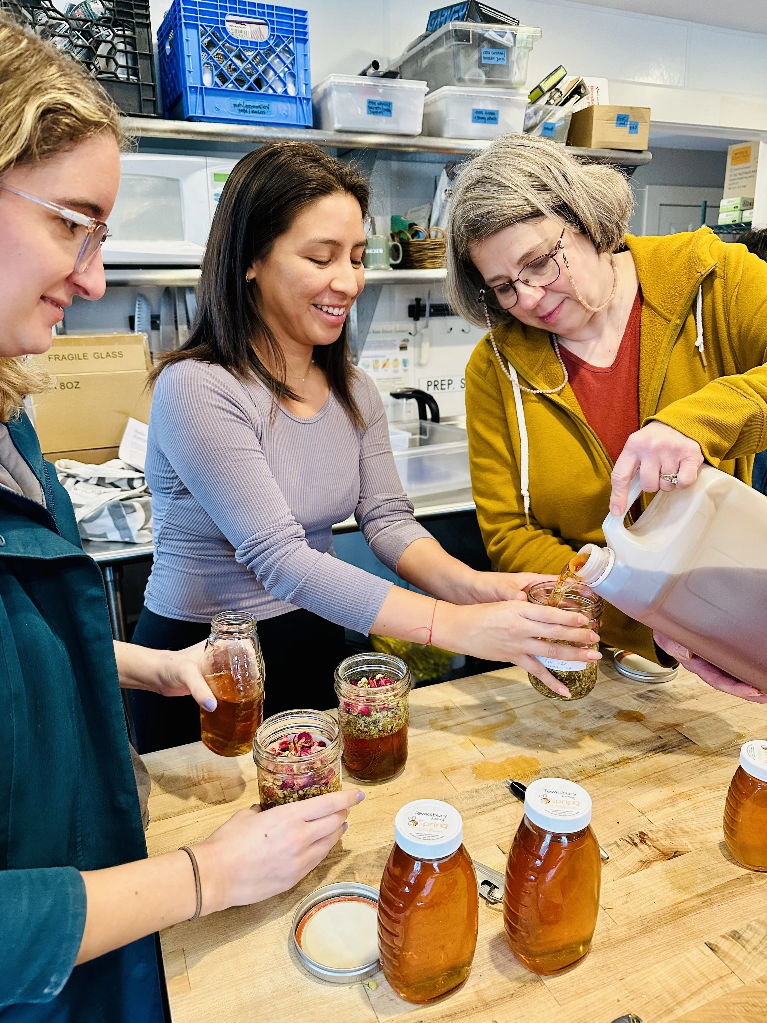Three women in a kitchen pouring honey into jars with dried flowers, surrounded by jars and supplies on the table.