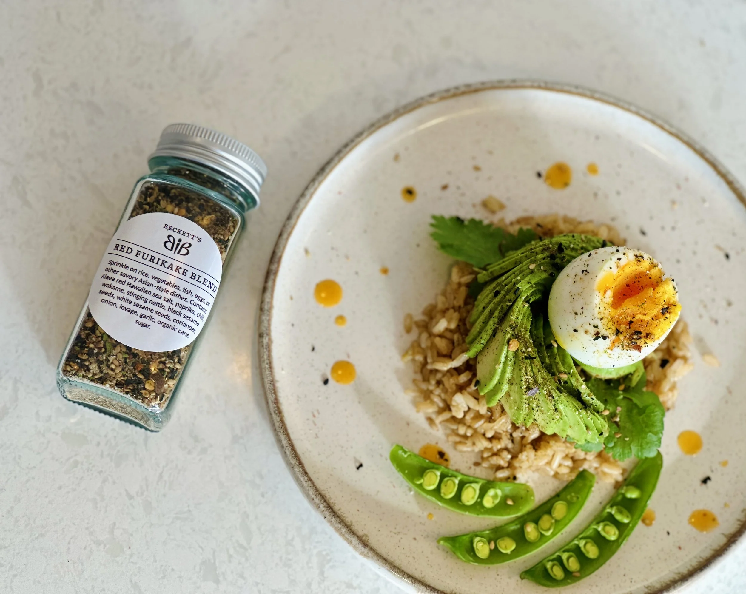 A plate with rice, sliced avocado, a halved soft-boiled egg, and snap peas, garnished with fresh herbs, a drizzle of oil, and colorful spice sprinkled on top. Next to the plate is a jar of red furikake seasoning.