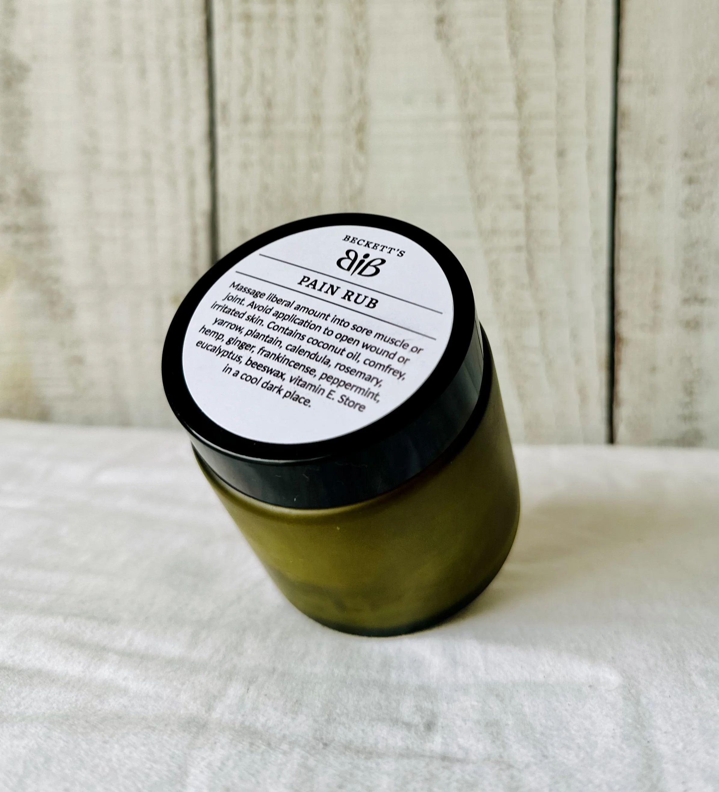 A jar labeled 'Beckett's Pain Rub' sitting on a white surface with a wooden background.