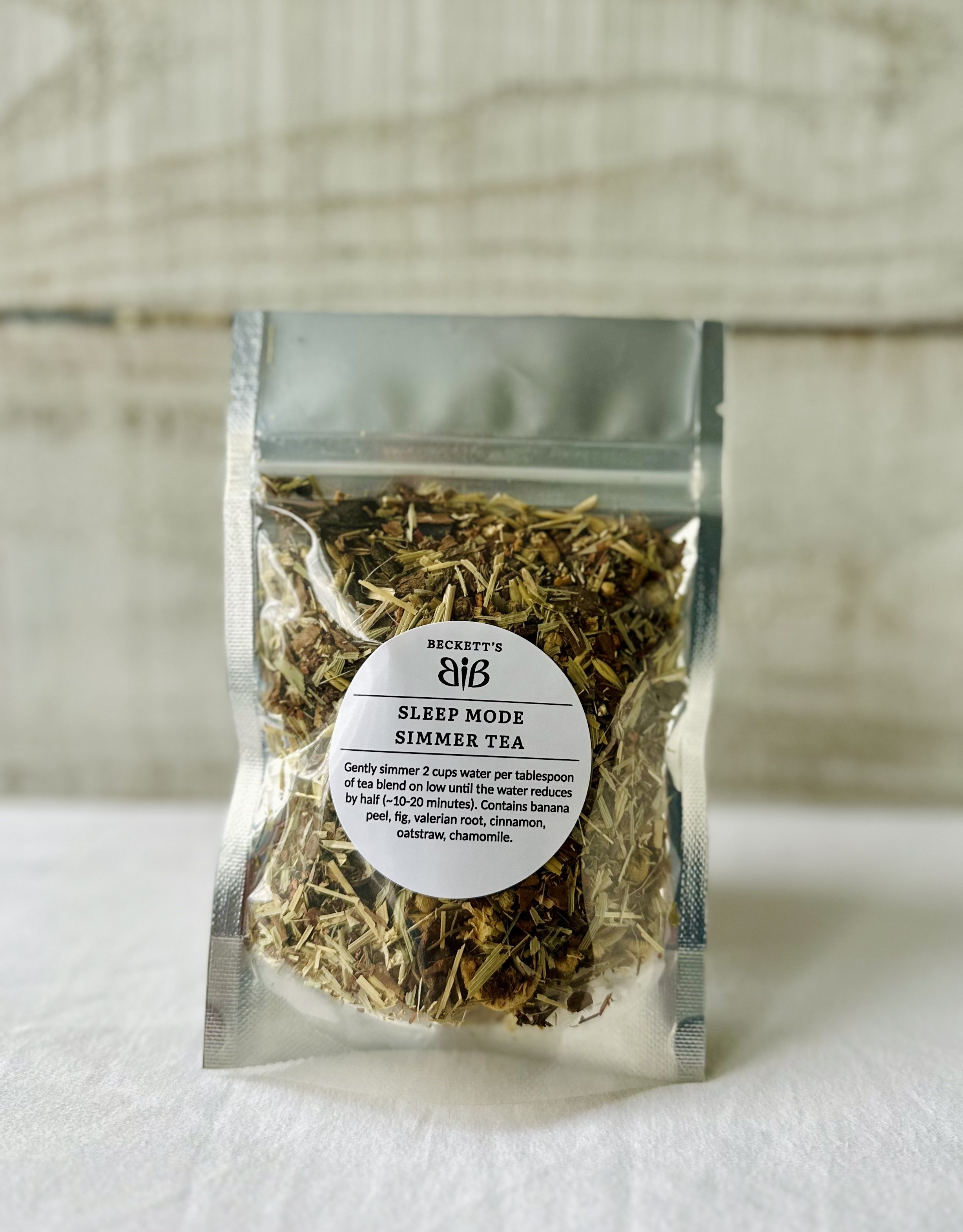 A clear resealable bag of herbal tea labeled 'Sleep Mode Simmer Tea' from Beckett's, placed on a white surface with a blurred wooden background. The tea contains ingredients like banana peel, fig, valerian root, cinnamon, oatstraw, and chamomile.