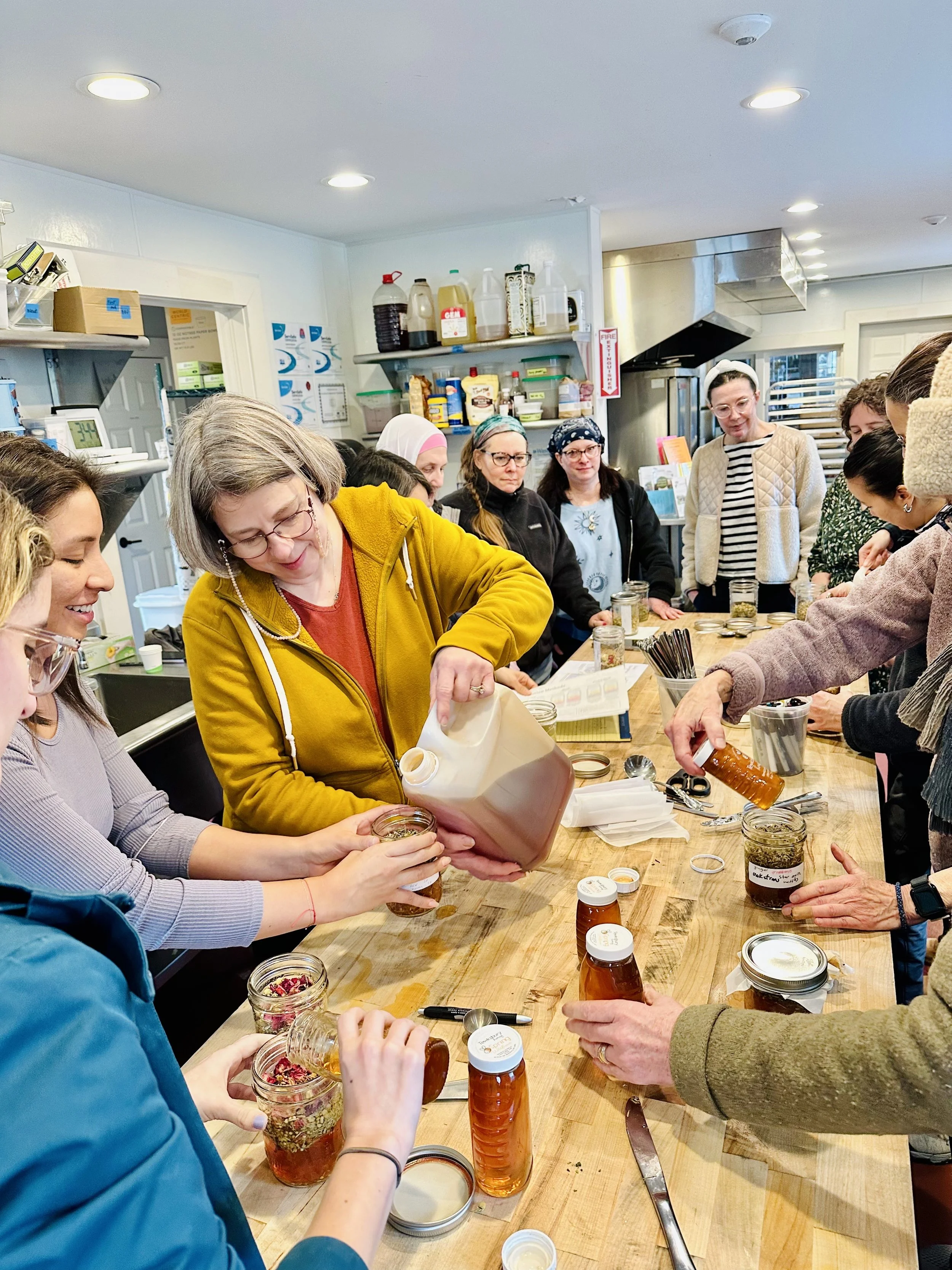Group of women gathered around a wooden table in a kitchen, pouring and preparing herbal vinegars in jars.