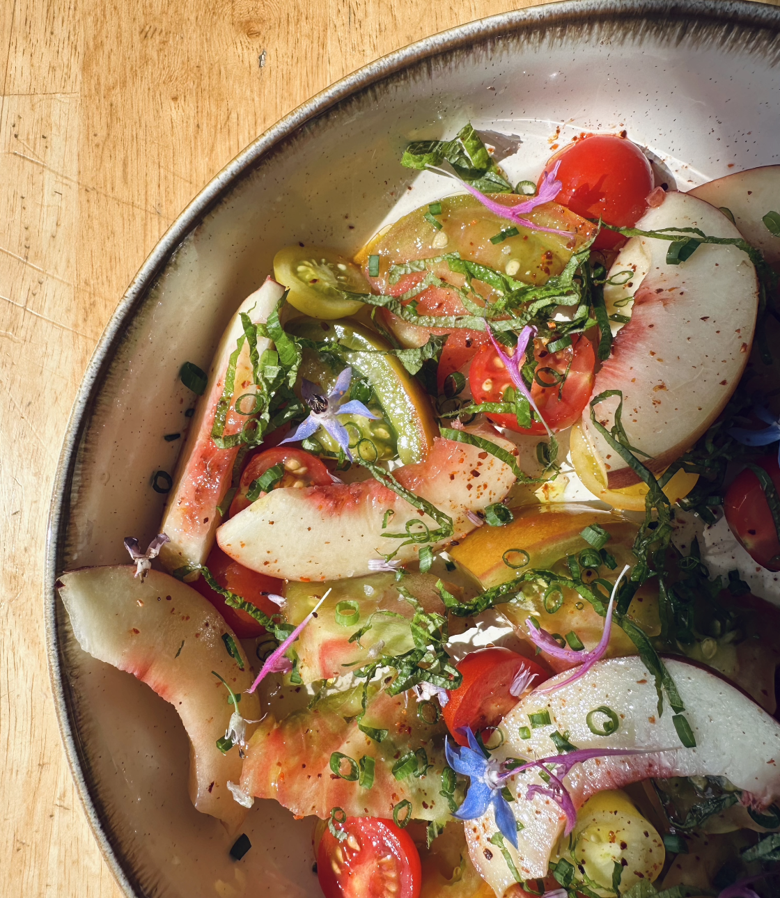 A colorful mixed vegetable salad with cherry tomatoes, sliced peaches, chopped herbs, and edible flowers in a ceramic bowl on a wooden surface.
