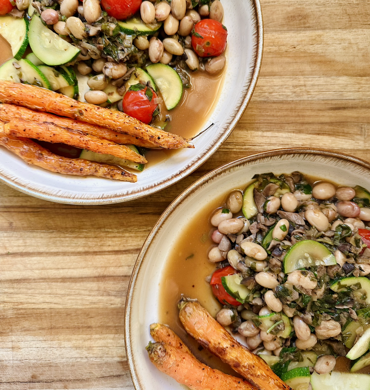 Two bowls of lentil and vegetable soup with cherry tomatoes and sliced zucchini, served with roasted carrots on a wooden surface.