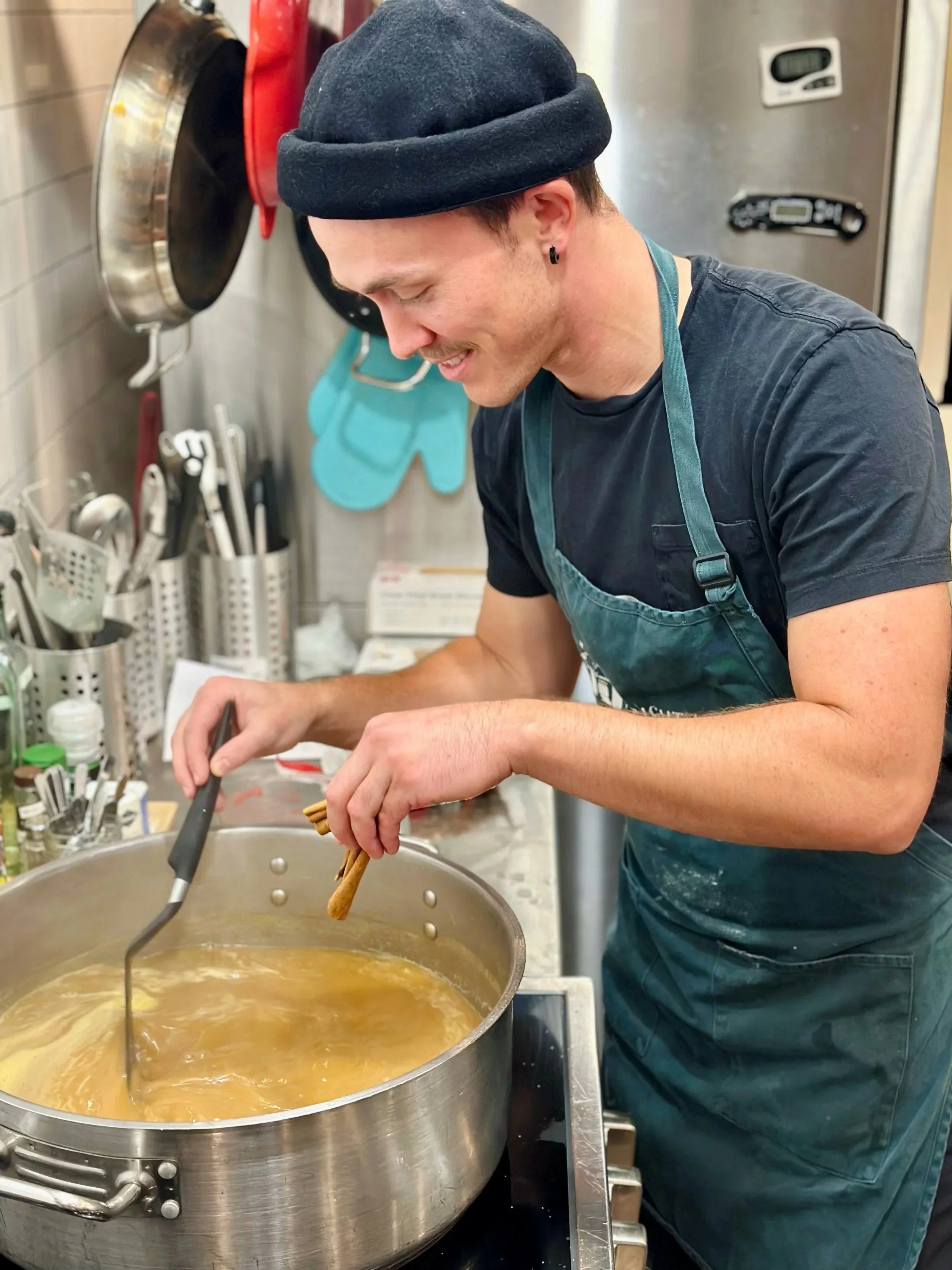 A smiling man wearing a black beanie, black t-shirt, and apron stirring a pot herbal plant-based milk tea on a stove in a kitchen.