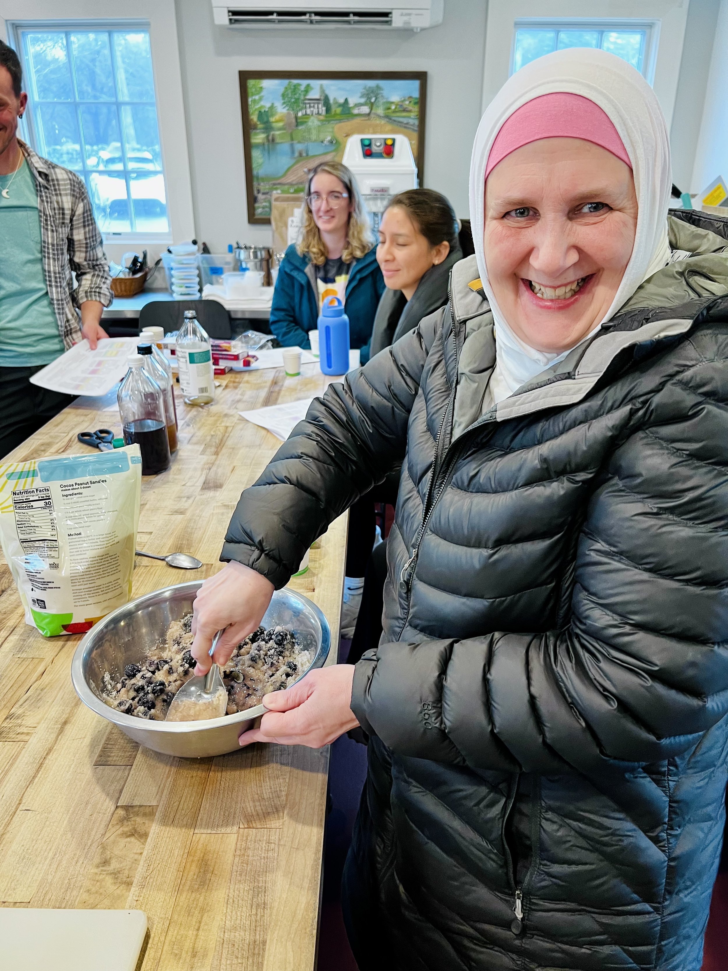 A smiling woman in a black puffy jacket and a white and pink headscarf mixing ingredients for an infused vinegar in a bowl. Other people are sitting at a table with ingredients and supplies in a bright room with windows.
