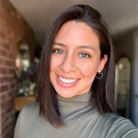 A young woman with shoulder-length dark hair, fair skin, and blue eyes smiling at the camera. She is wearing a gray turtleneck sweater and small hoop earrings, standing indoors with a blurred background.