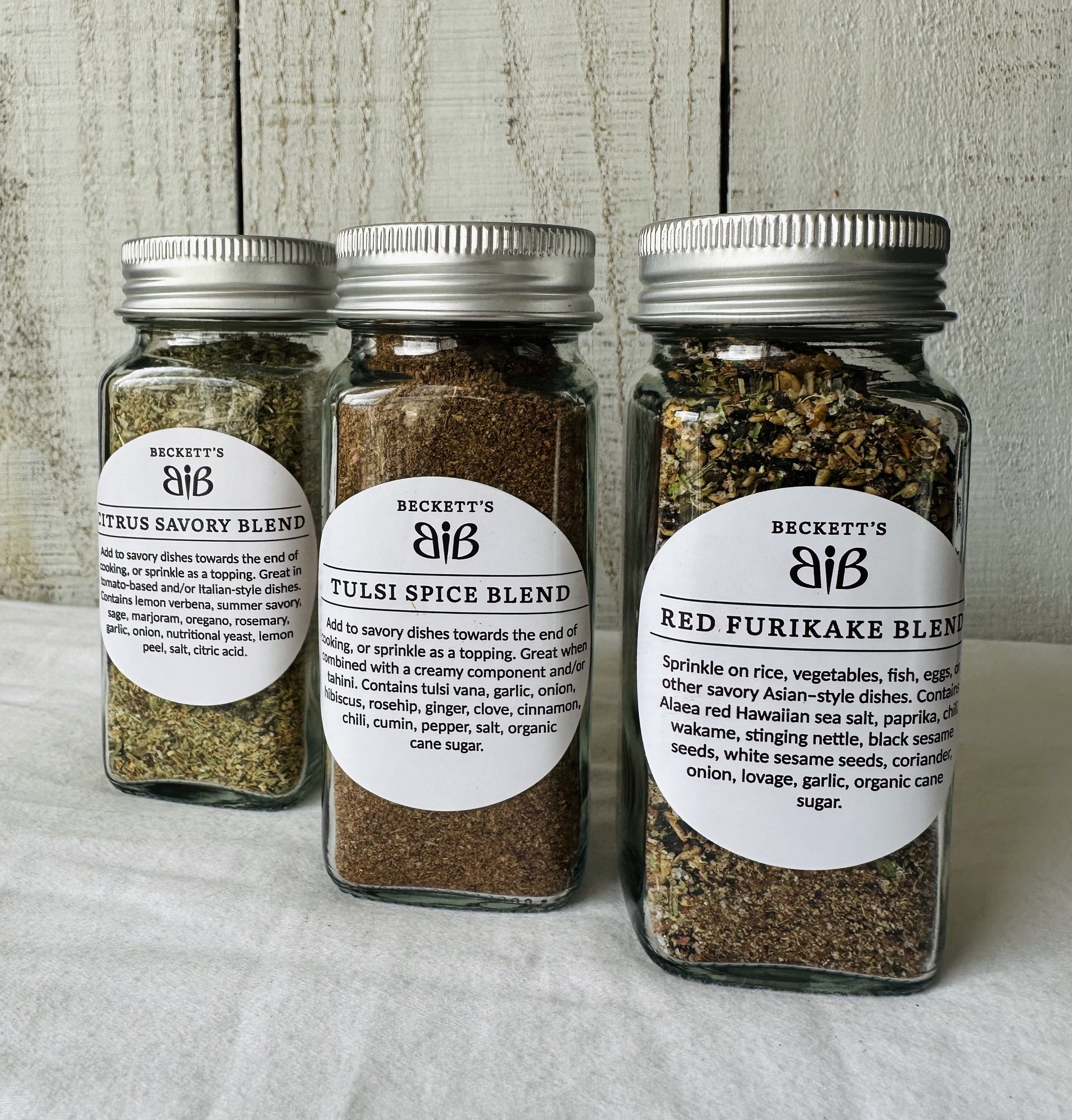 Three glass jars with metal lids containing various spice blends, labeled from left to right as Citrus Savory Blend, Tulsi Spice Blend, and Red Furikake Blend, with white labels and black text.