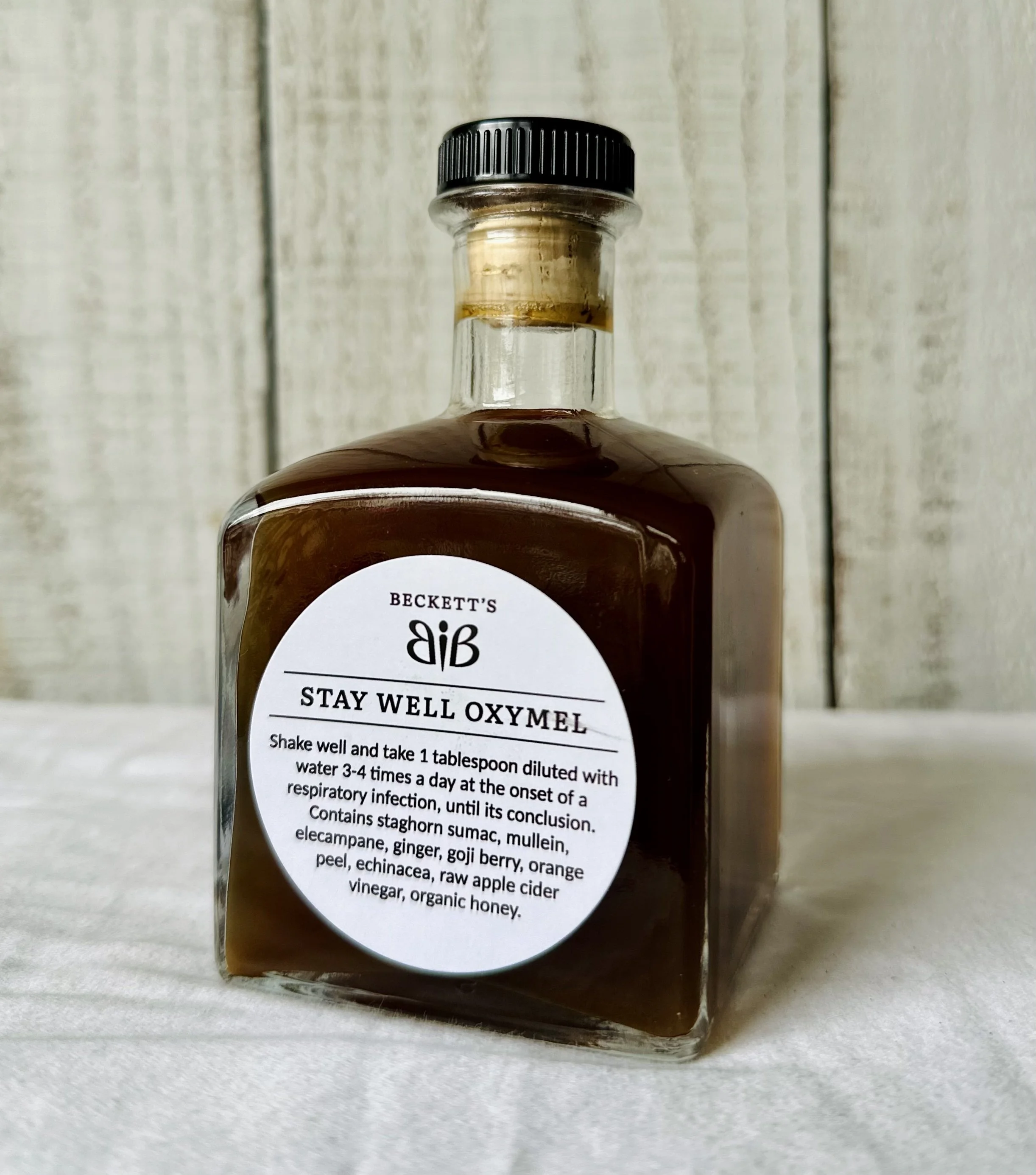 A glass bottle with a black cap containing a medicinal oxymel labeled 'Beckett's Stay Well Oxymel,' with instructions and ingredients on the label.