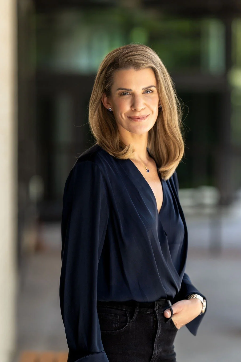 A woman with shoulder-length light brown hair smiling, wearing a dark blue blouse and black jeans, standing outdoors with a blurred background.