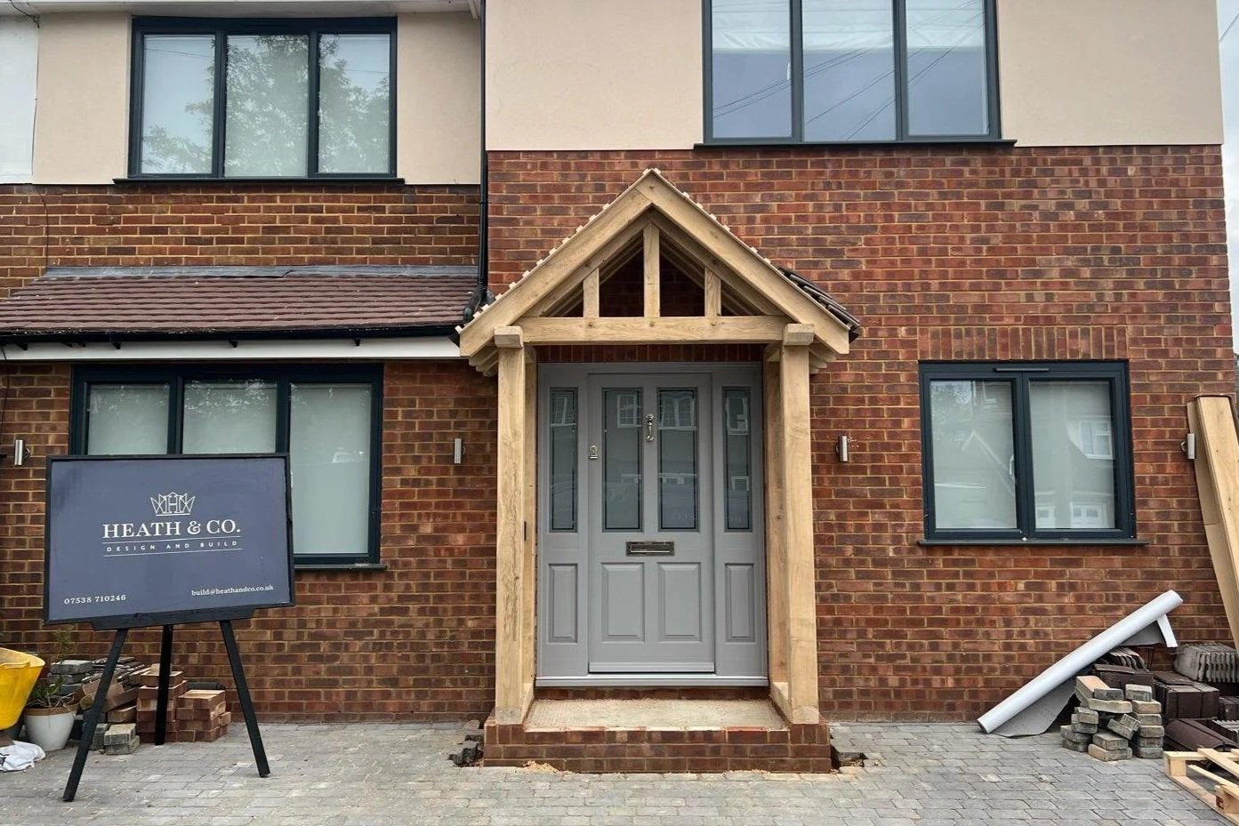 Newly constructed brick house with a gray front door under a small gabled porch, and four large windows with black frames on the upper and lower floors. A sign for HEATH & CO. Design and Build is on the left, and construction materials are on the right.