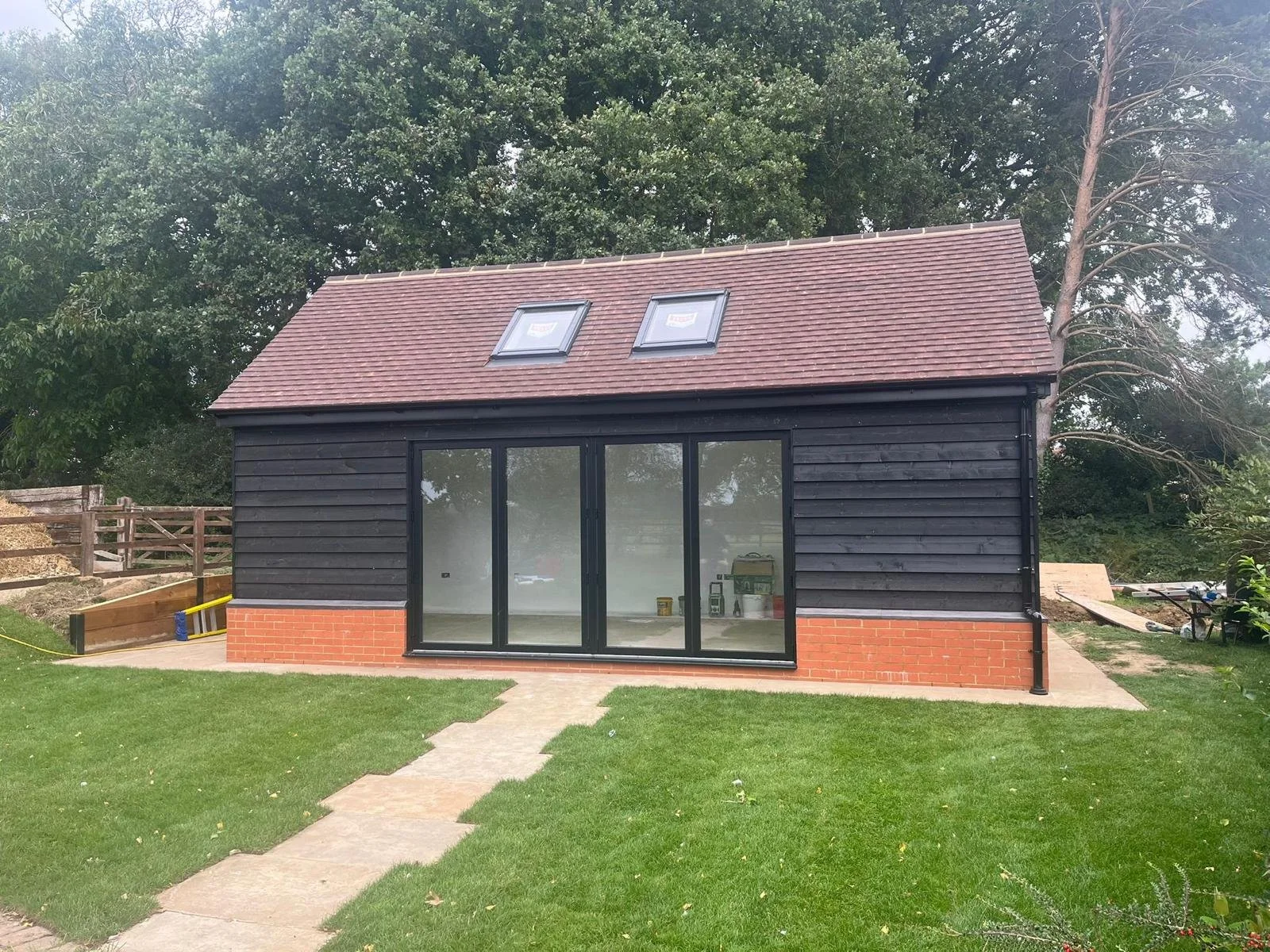 Newly constructed black wooden garden shed with brick foundation, large glass sliding door, and two skylights on the roof, situated in a grassy backyard with trees in the background.