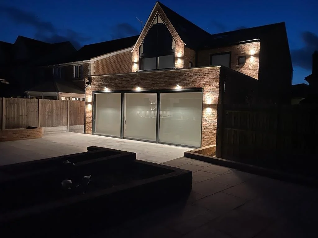 A two-story brick house at night with outdoor lighting on the back patio, featuring large glass sliding doors, a wooden fence, and a paved yard.