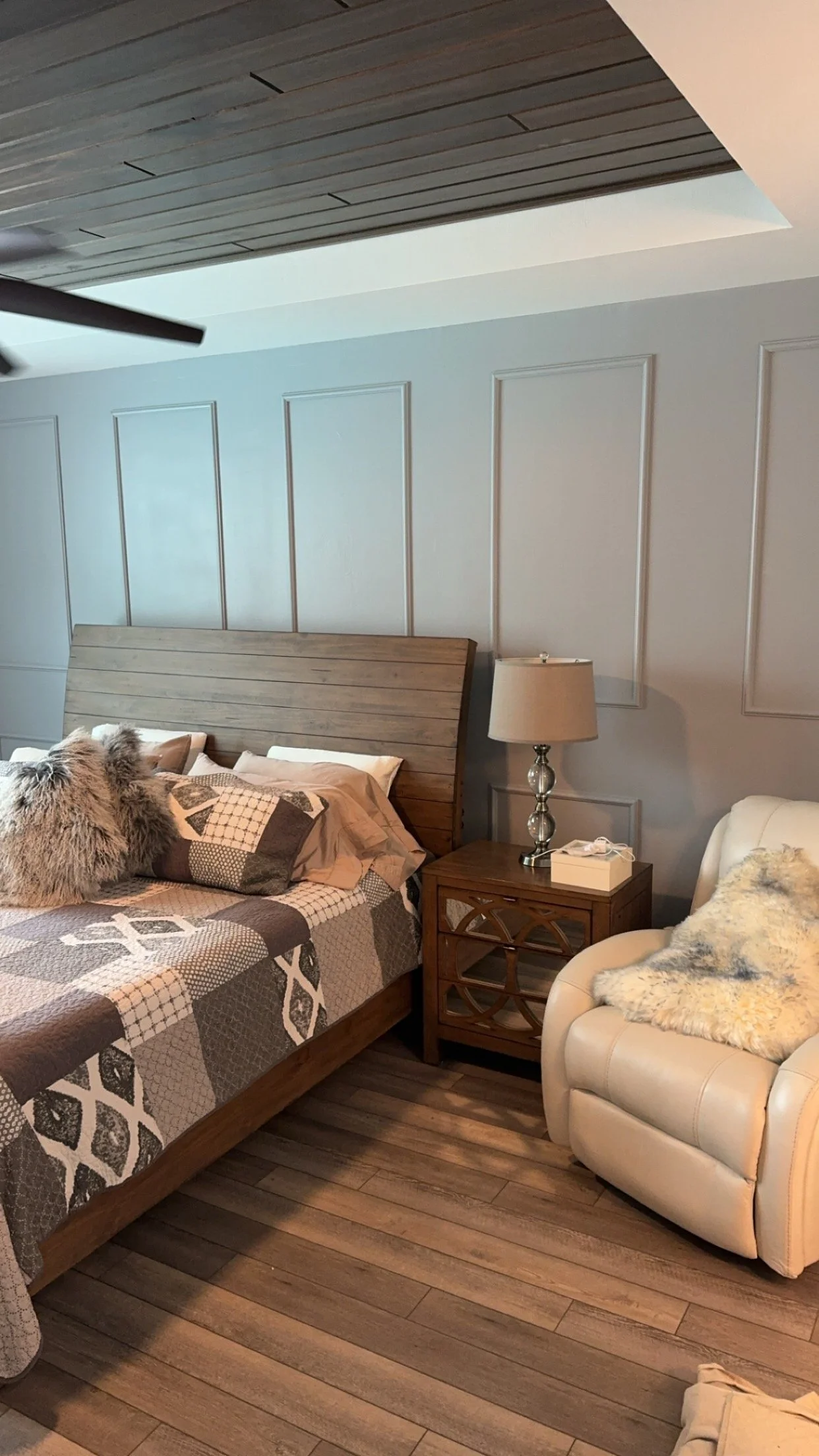 Bedroom with wooden bed frame, quilted bedspread, fluffy pillow, wooden nightstand with lamp, cream recliner with furry blanket, wooden flooring, and molding on the wall. Trim accent wall. Painted accent wall.