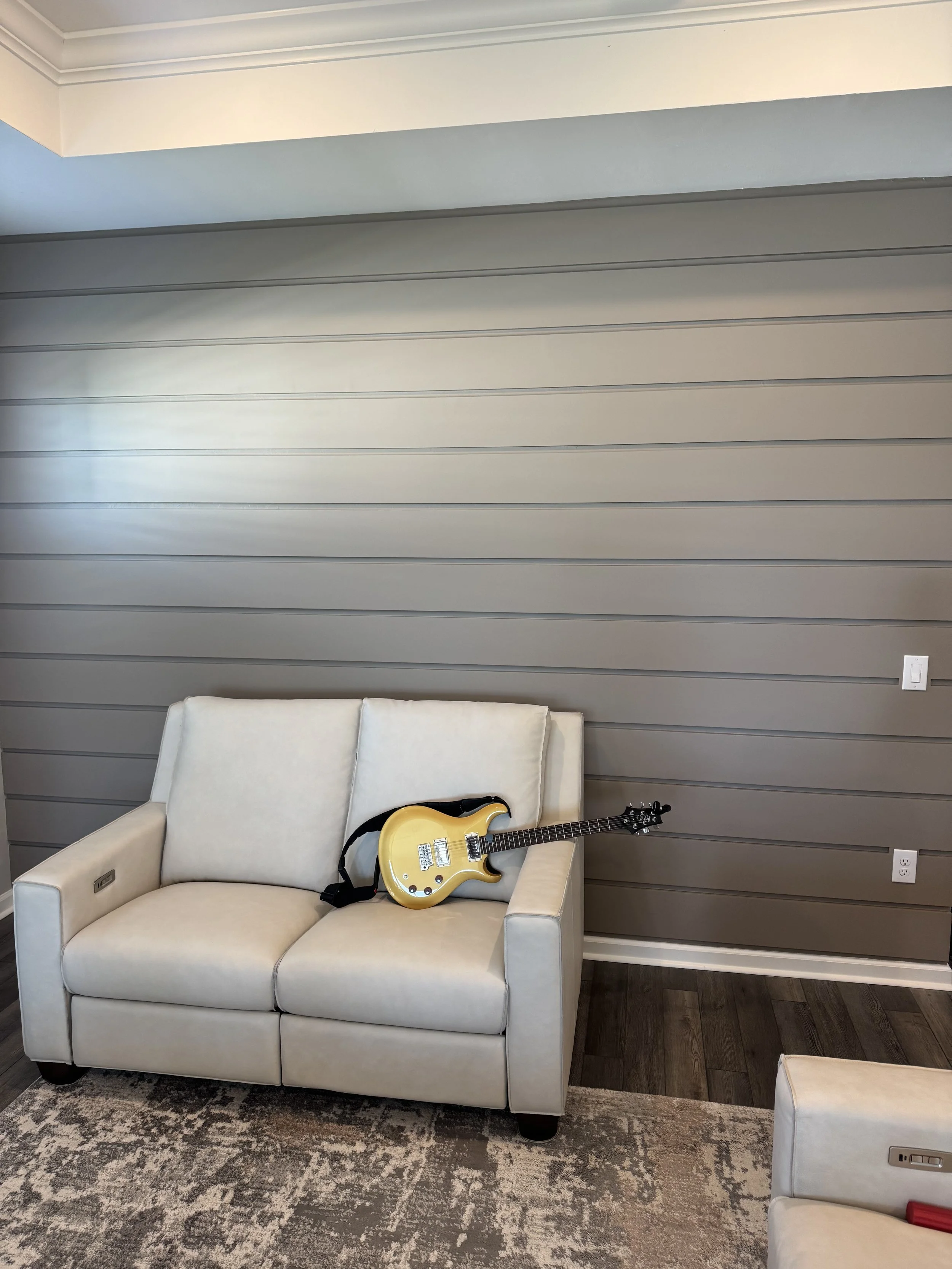 A yellow electric guitar resting on the arm of a white sofa in a living room with a wall featuring horizontal panels.