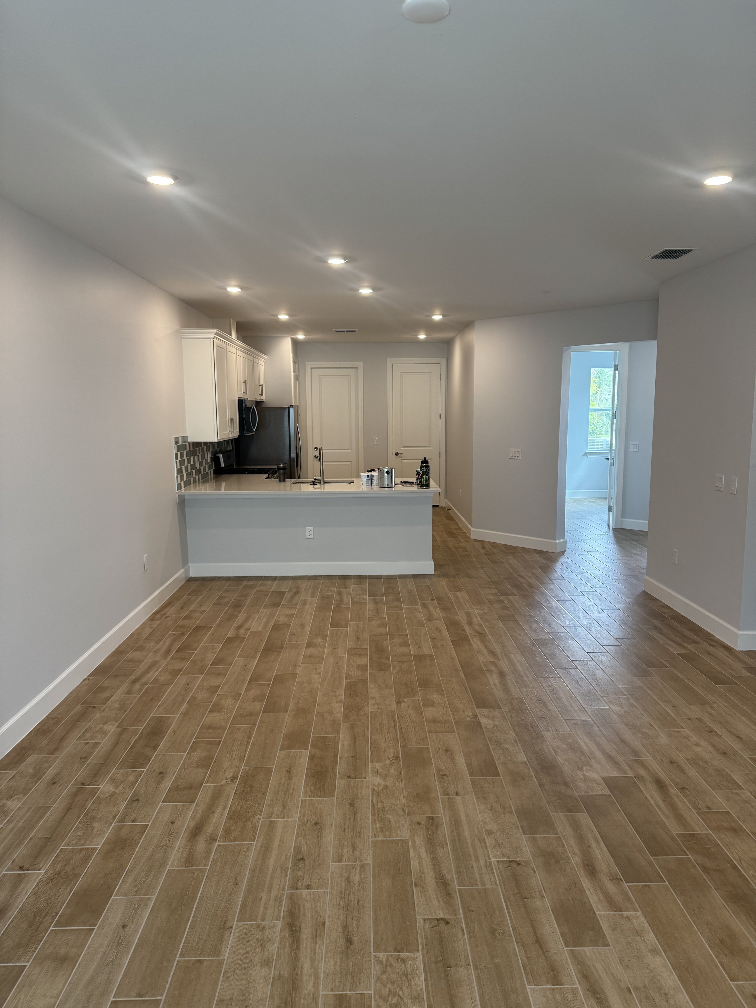 Empty modern kitchen and living area with wood-look tile flooring, white walls, recessed lighting, and a small breakfast bar.