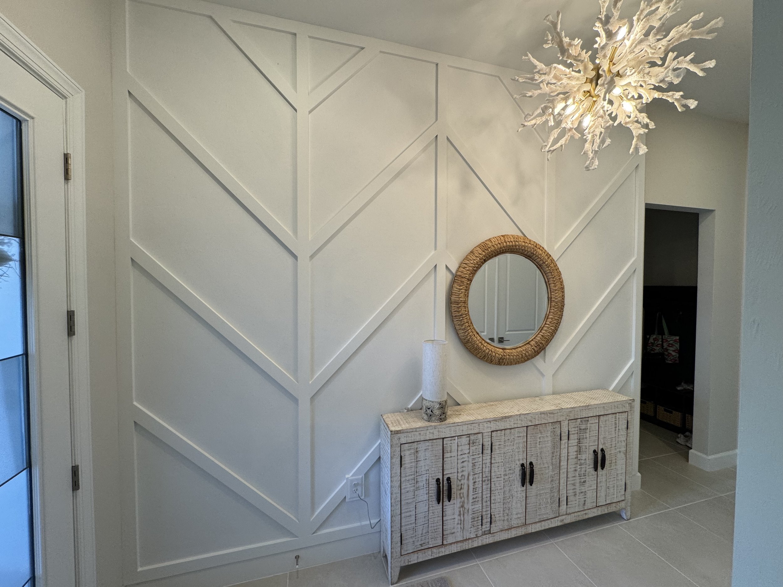A modern entryway with a textured white wall, a round wicker mirror, a rustic white sideboard, a white decorative lamp, and a chandelier resembling coral.