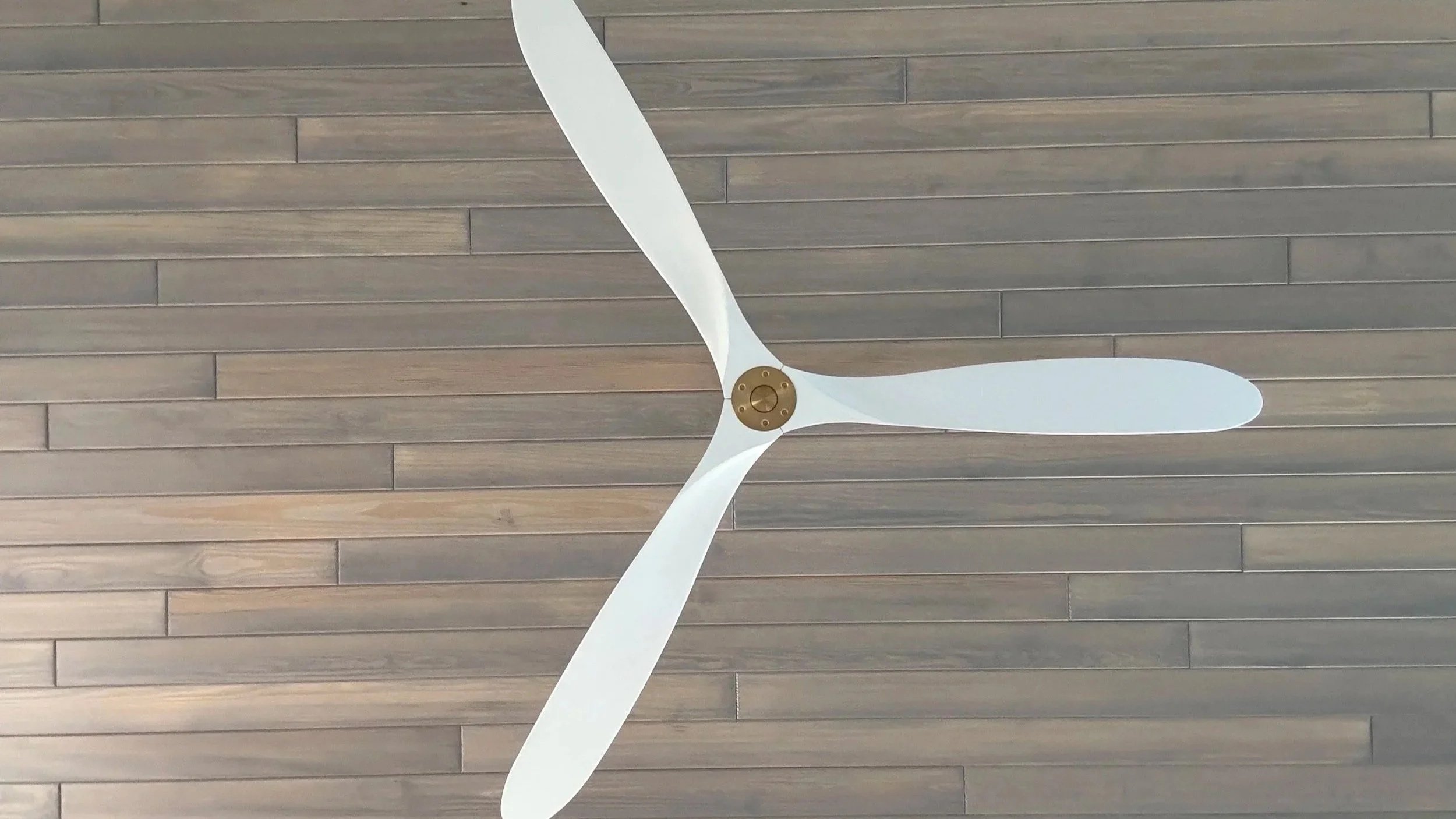 Close-up of white ceiling fan with three blades mounted on a brown wooden ceiling. Tongue and Groove ceiling