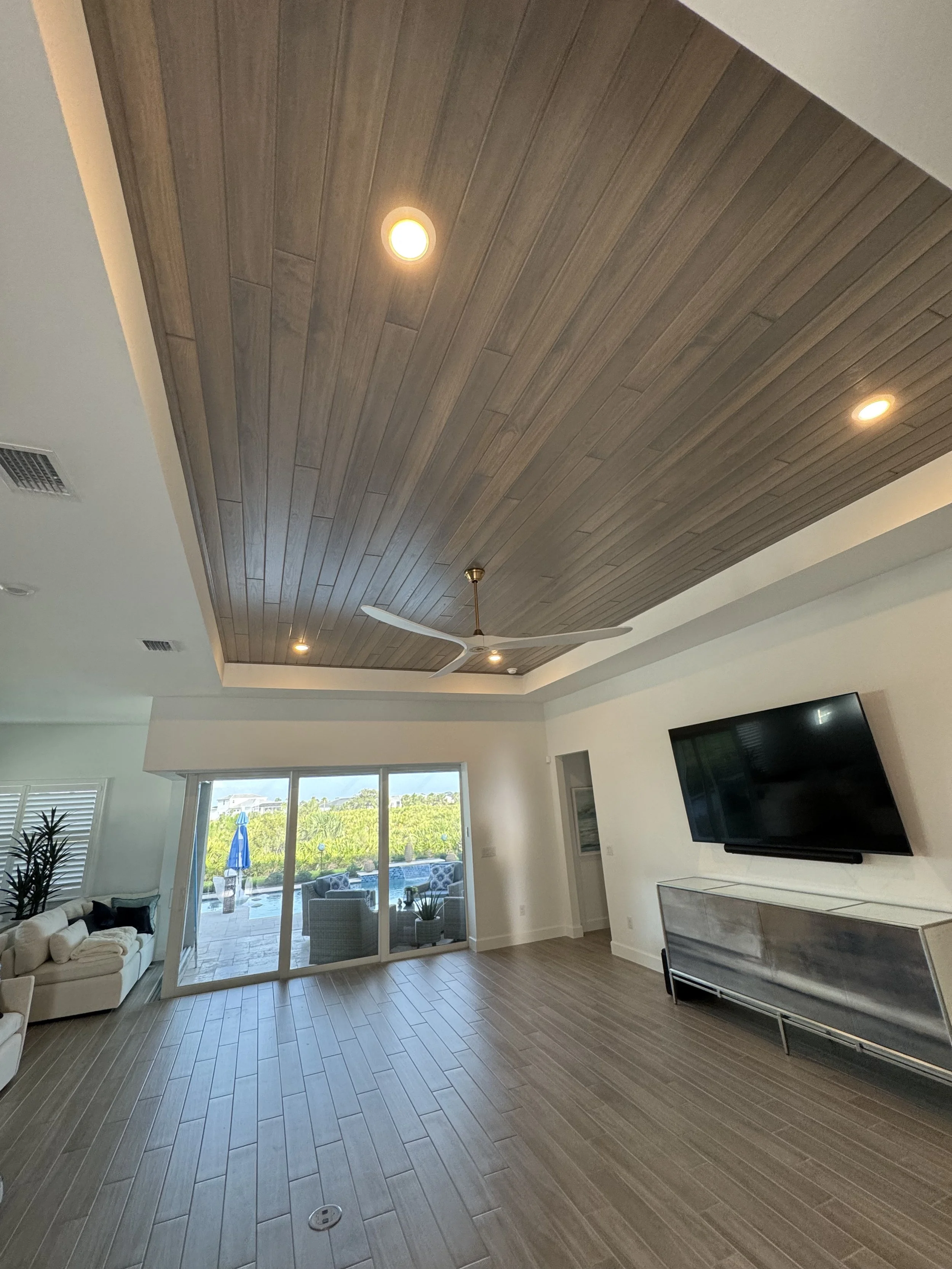 Living room with wooden ceiling, ceiling fan, recessed lighting, sliding glass door leading to outdoor patio with pool, sofa, and chairs, TV on white media console. Tongue and Groove ceiling.