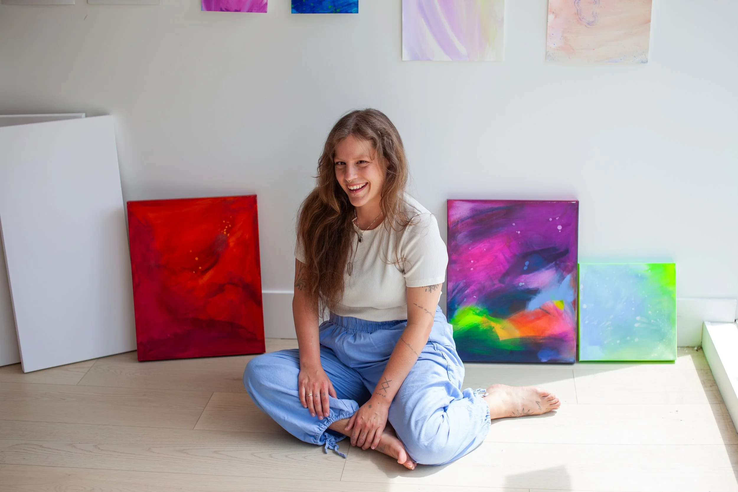 A woman with long wavy hair sitting on a light wooden floor, smiling, surrounded by colorful abstract paintings leaning against a white wall.