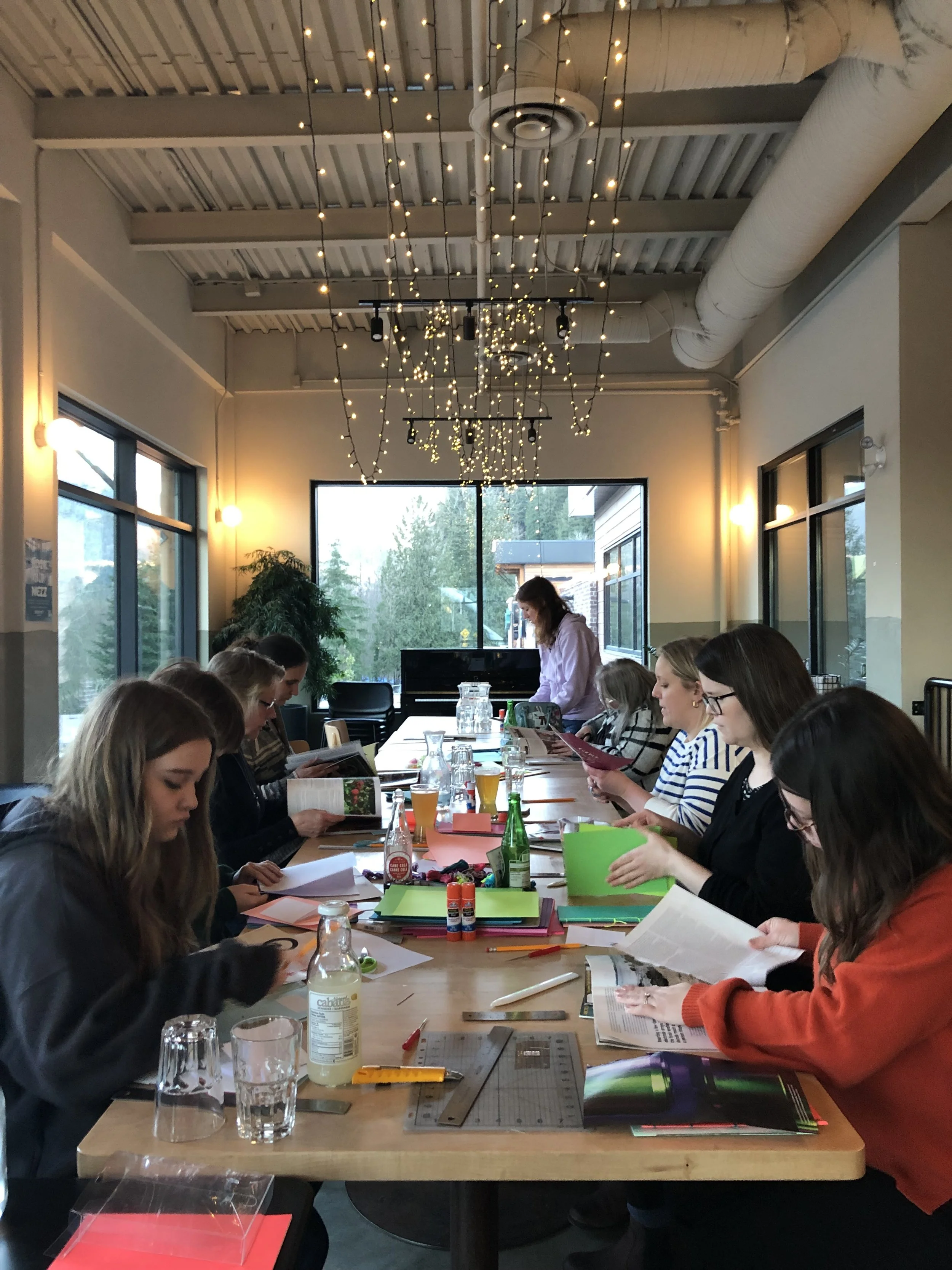 People sitting around a long table in a brightly lit room with large windows, plants, and decorative string lights on the ceiling, engaged in reading or writing activities.