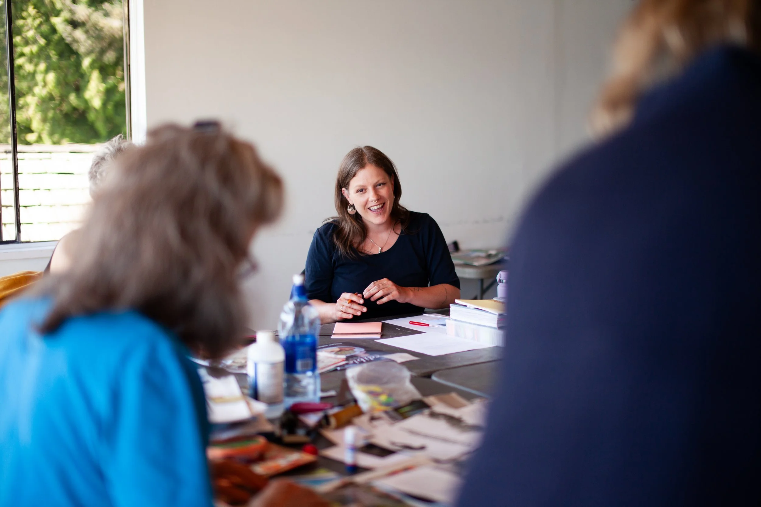 A woman sitting at a table, smiling and engaging with others during a discussion or meeting, with art supplies and papers on the table.