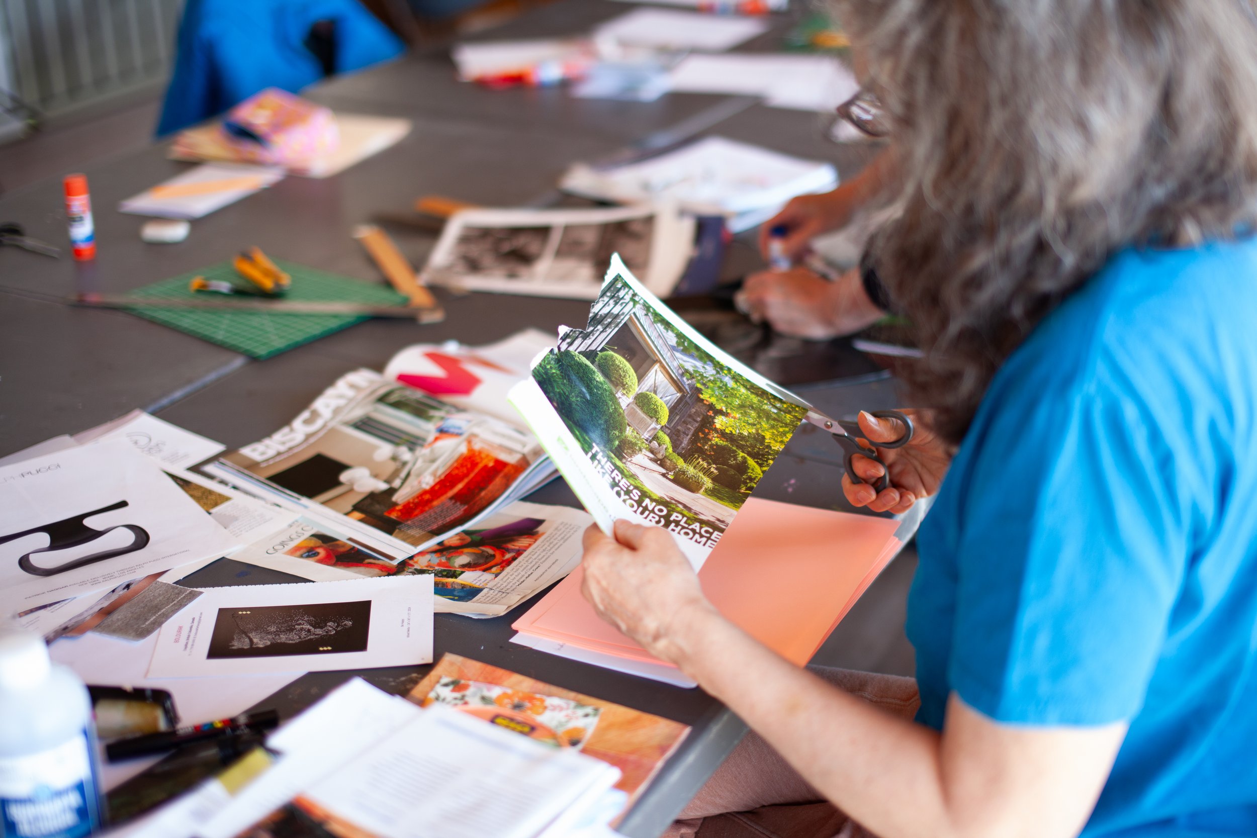 Person with long curly hair wearing a blue shirt cutting a magazine with scissors at a cluttered table surrounded by papers, magazines, and craft supplies.